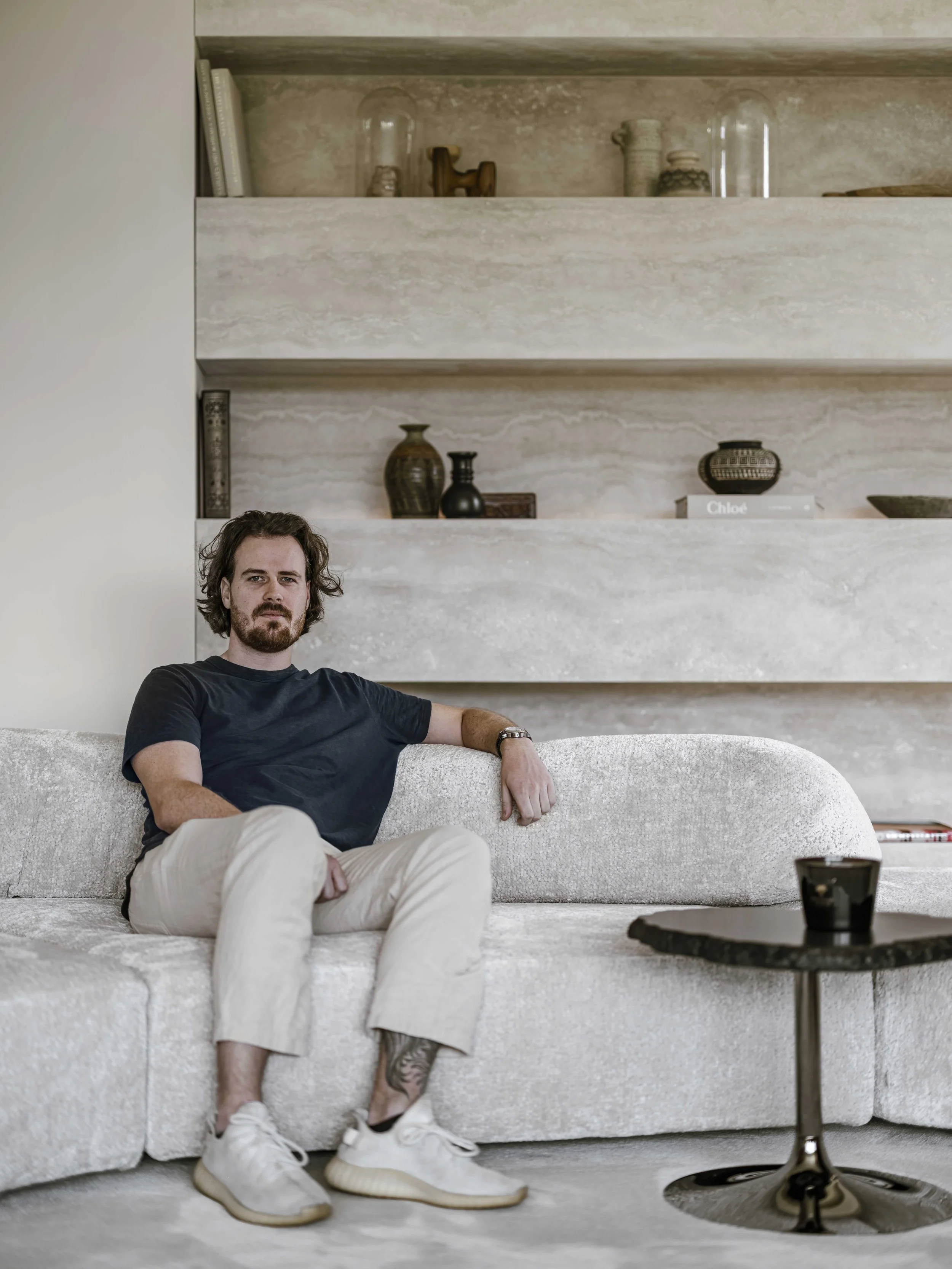 Guy de Vos, a man with dark hair, a beard, and tattoos on his leg sitting on a light Edra sofa in a modern living room with beige and neutral decor.