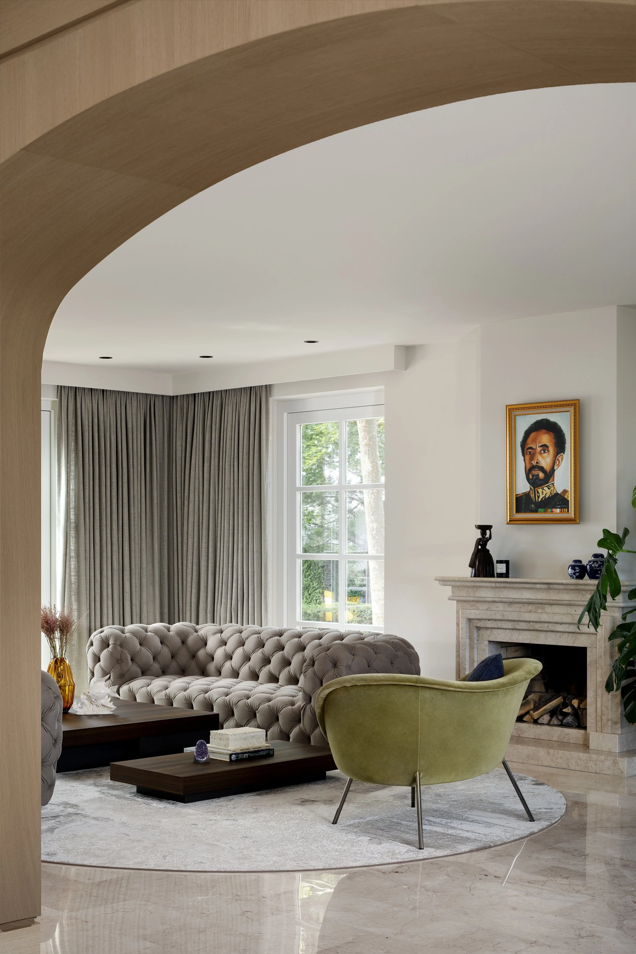Living room with a beige tufted Baxter sofa, green armchair, marble fireplace with artwork above, large window with curtains, and decorative items on the mantel.