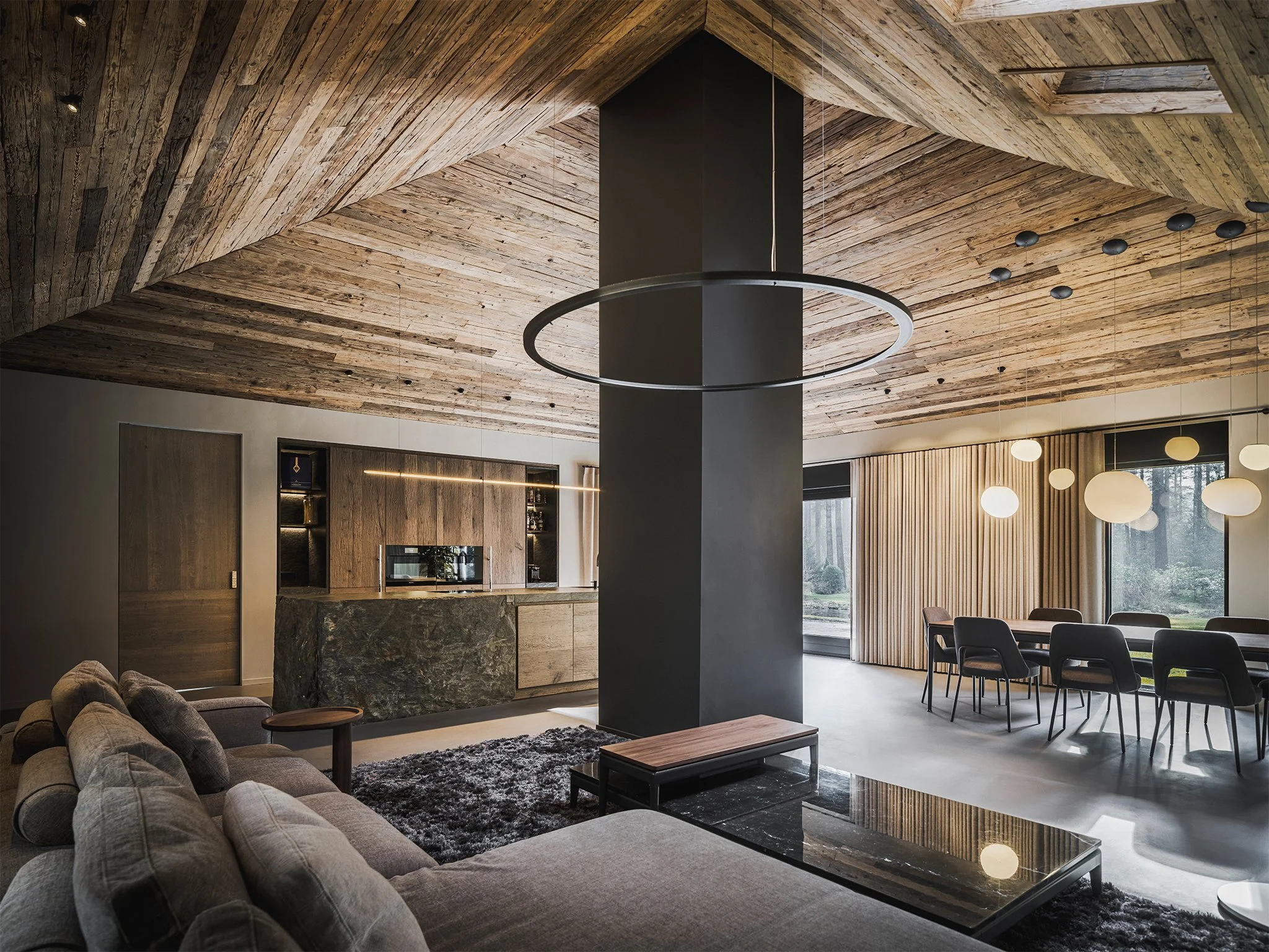 Chalet style living room with wooden ceiling, black pillar in center, gray Flexform sofa, coffee table, and a dining area with circular hanging lights.