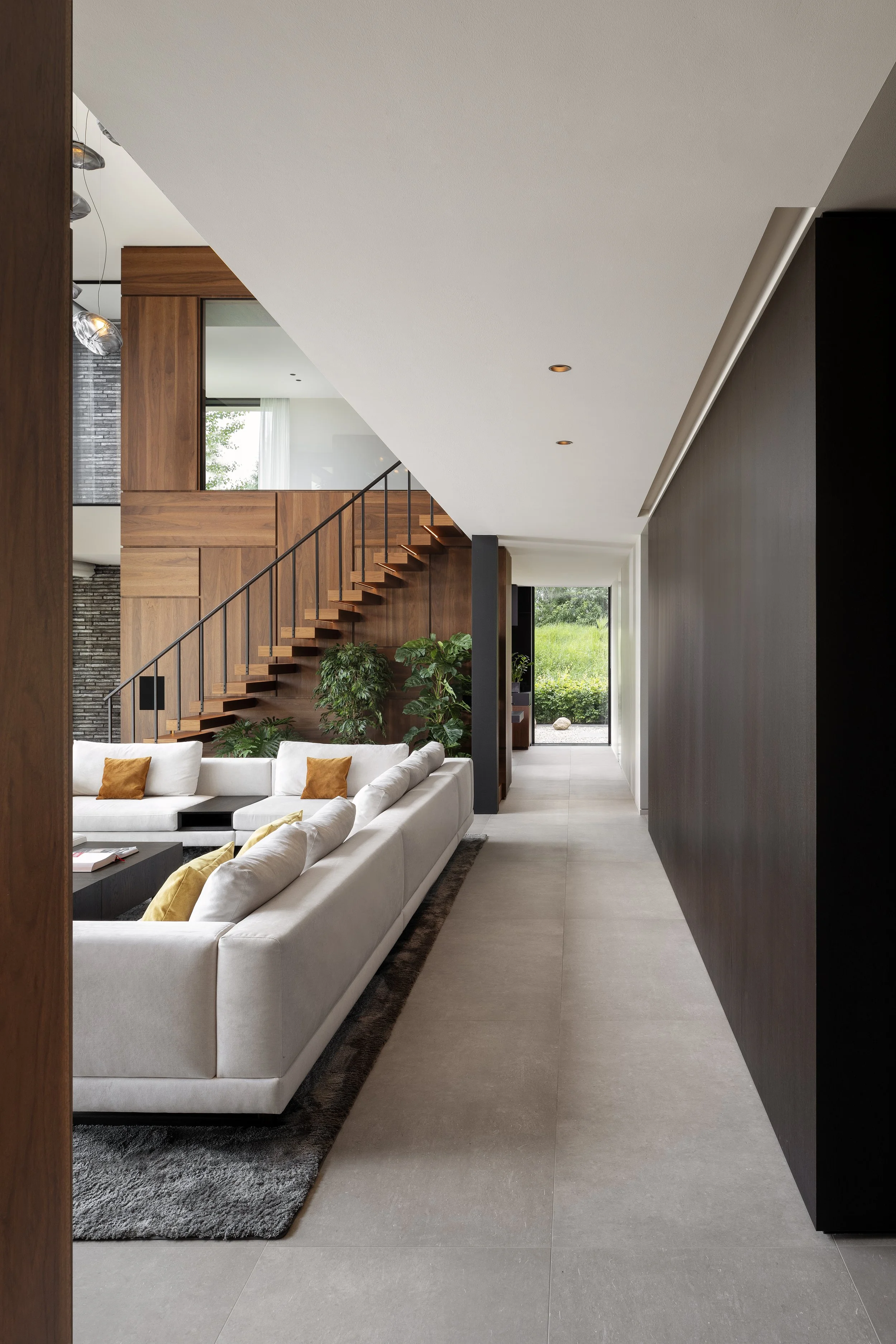 High quality designed living room with white sectional sofa, orange and yellow pillows, black coffee table, green indoor plants, wooden staircase with black railing, large glass door leading outside, beige tiled floor, dark wall on the right.