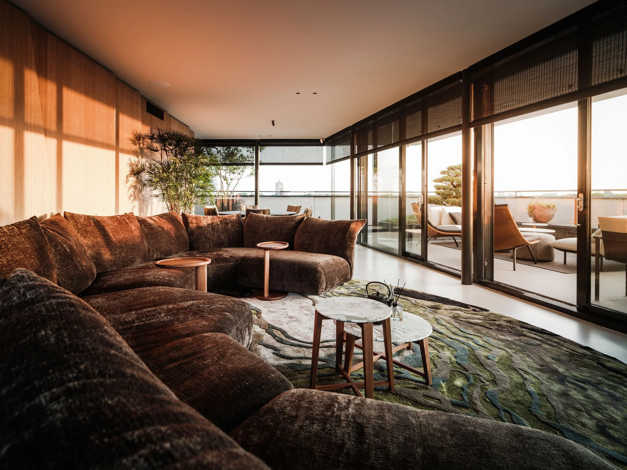 Luxurious living room with dark Edra sectional sofa, marble coffee tables, and large glass sliding doors leading to balcony with outdoor seating and potted plants, sunlit through the windows.