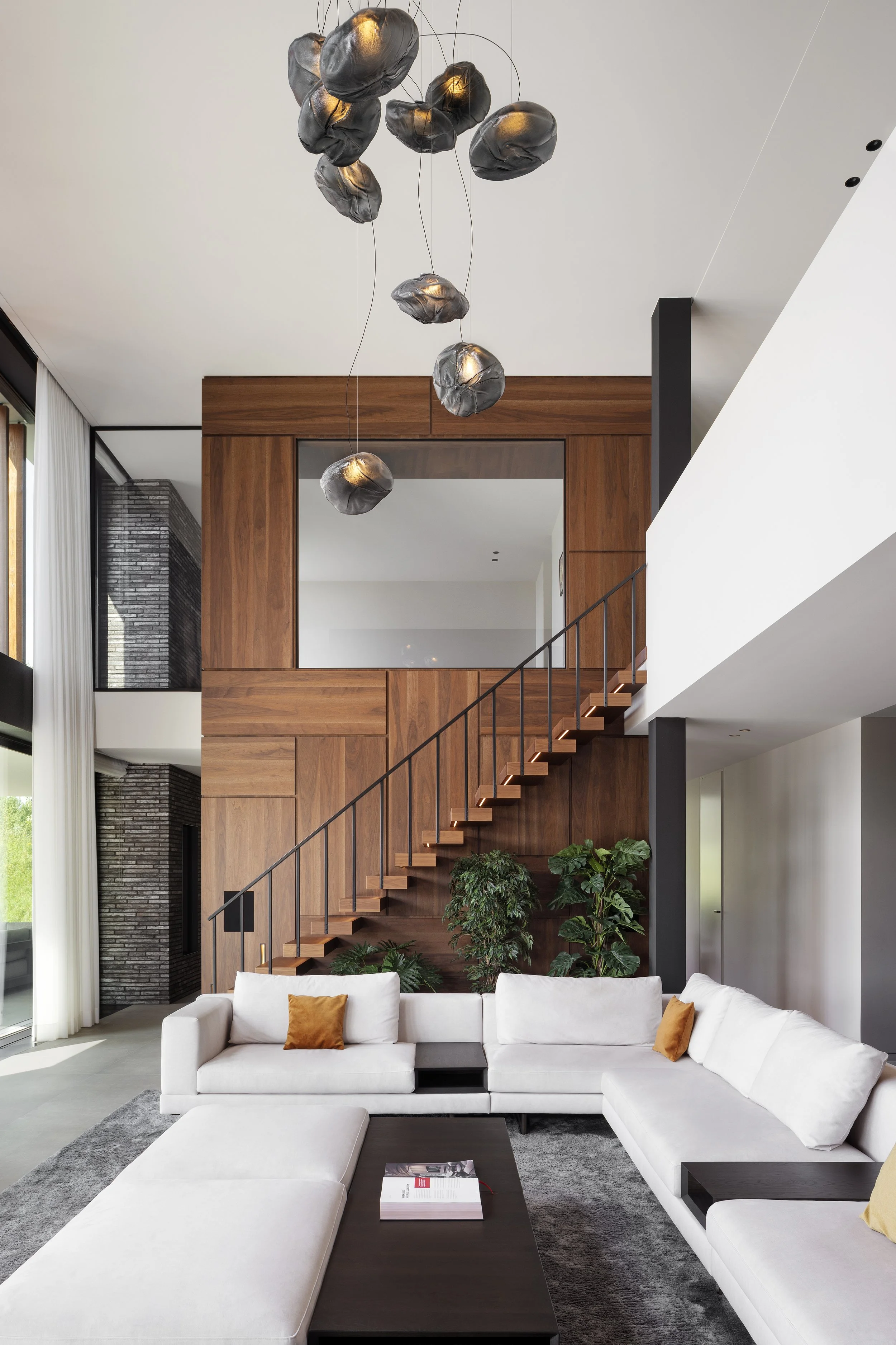 Double height living room with white sectional sofa, orange pillows, black coffee table with a magazine, green plants at the back, wooden staircase, and unique cloud-shaped Bocci pendant lights hanging from the ceiling.