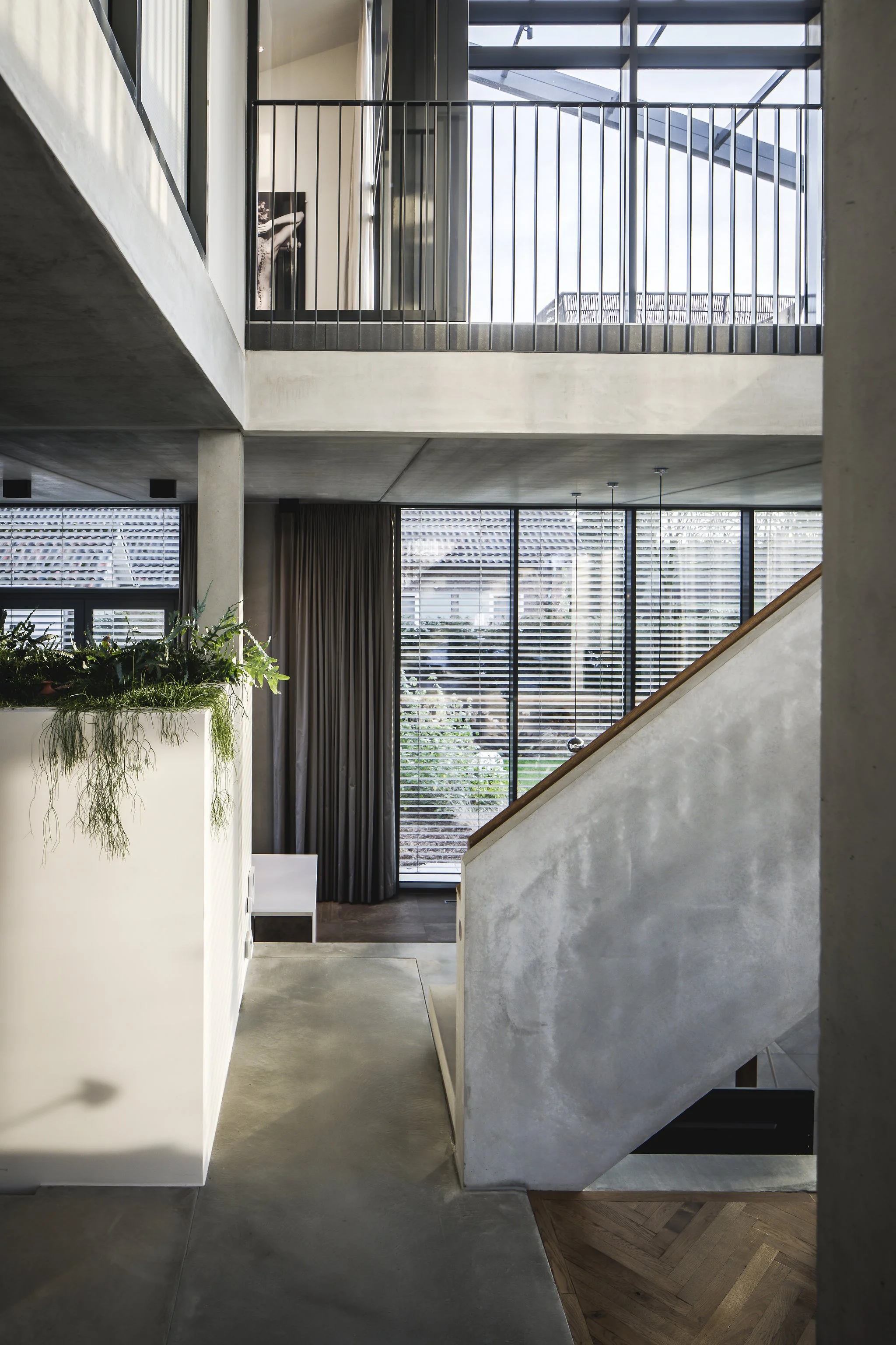 Interior view of a modern loft styled multi-story home with concrete stairs, large windows with horizontal blinds, glass door with outdoor stairs, and a balcony with a chair.