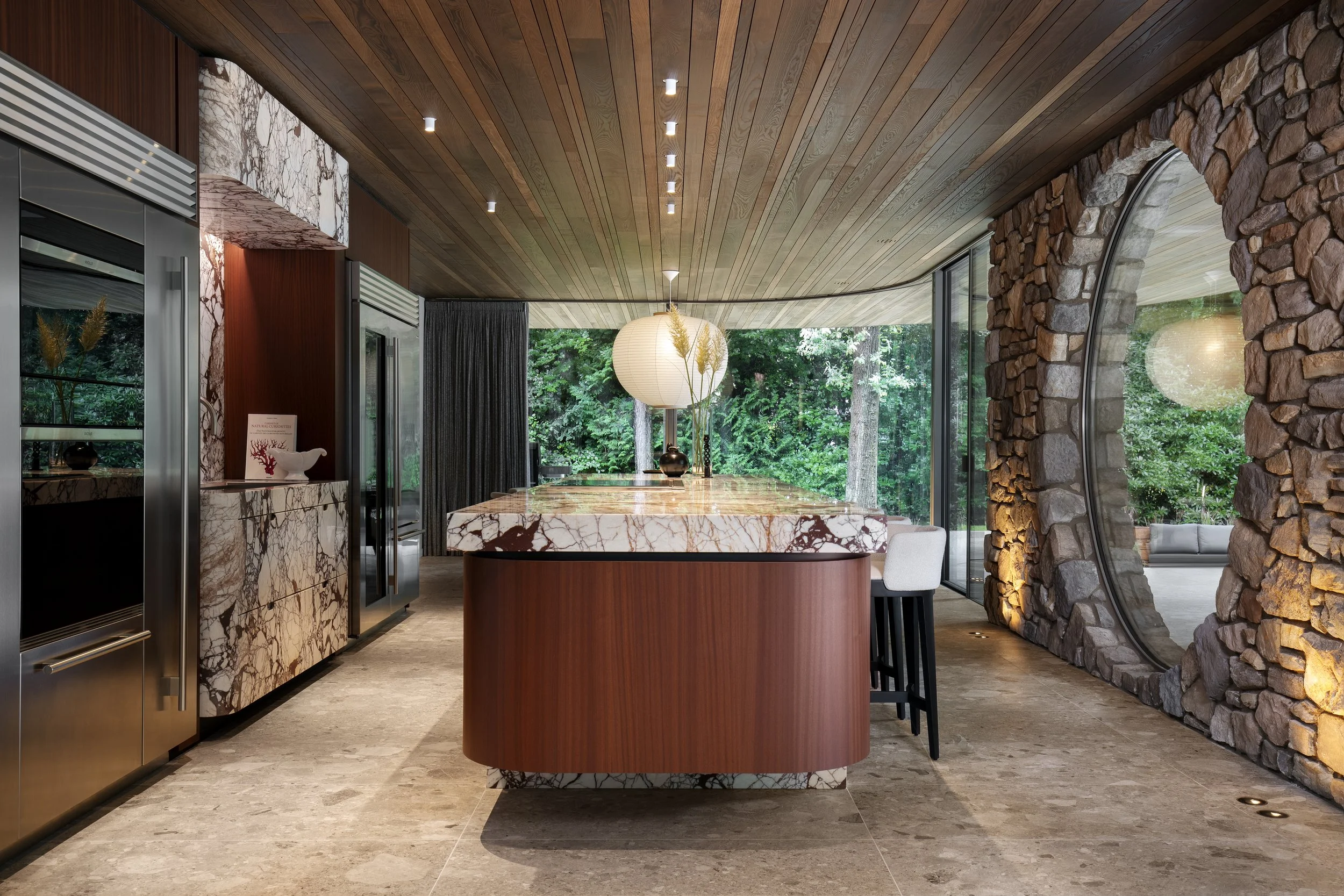 Modern kitchen with marble and wood island, subzero & wolf appliances, stone wall, and large round window showing trees outside.