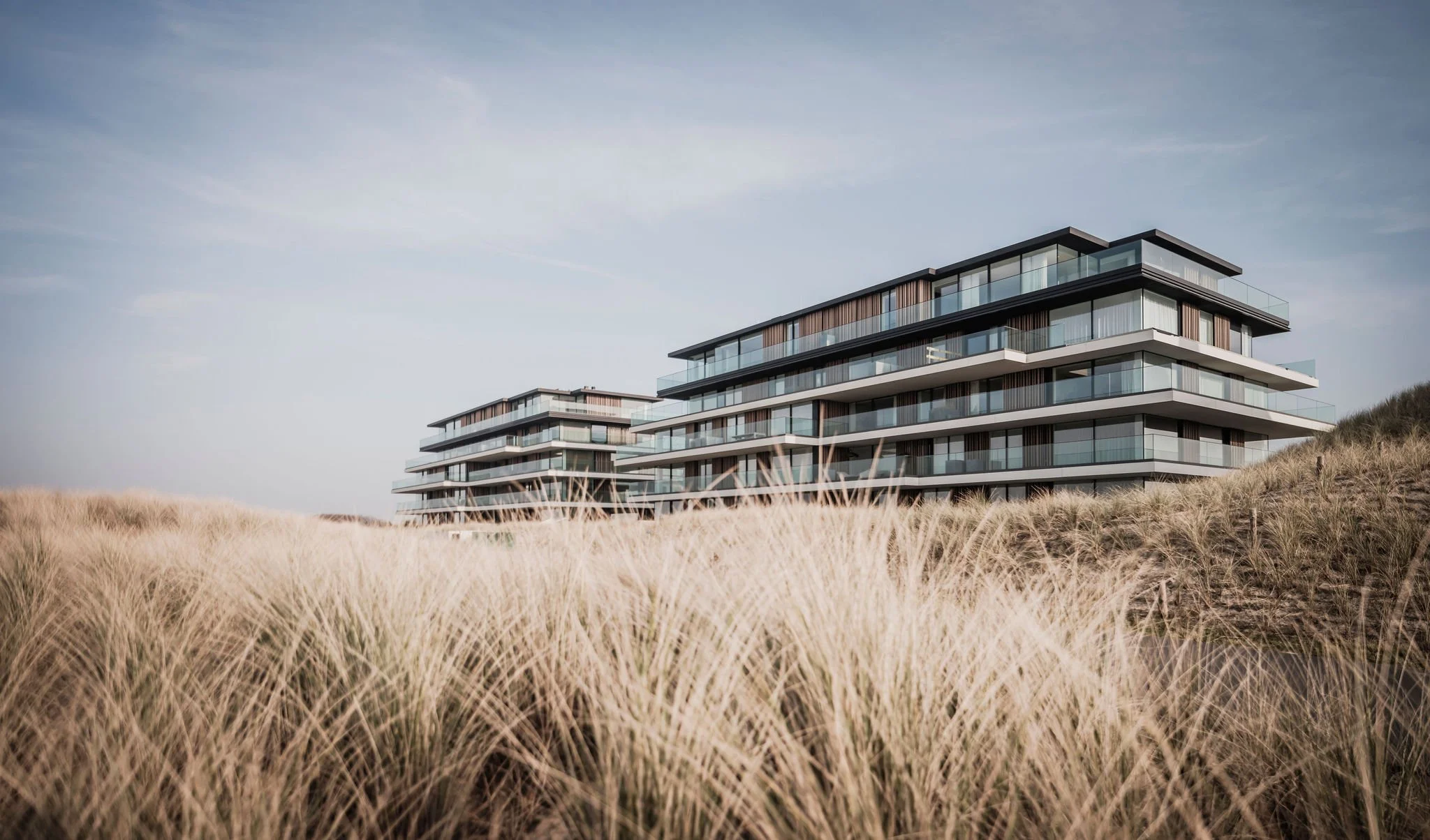 Modern multi-story ultra luxury apartment buildings with glass balconies situated behind a field of tall, dry grass, under a lightly clouded sky. located on the dunes overlooking the ocean.