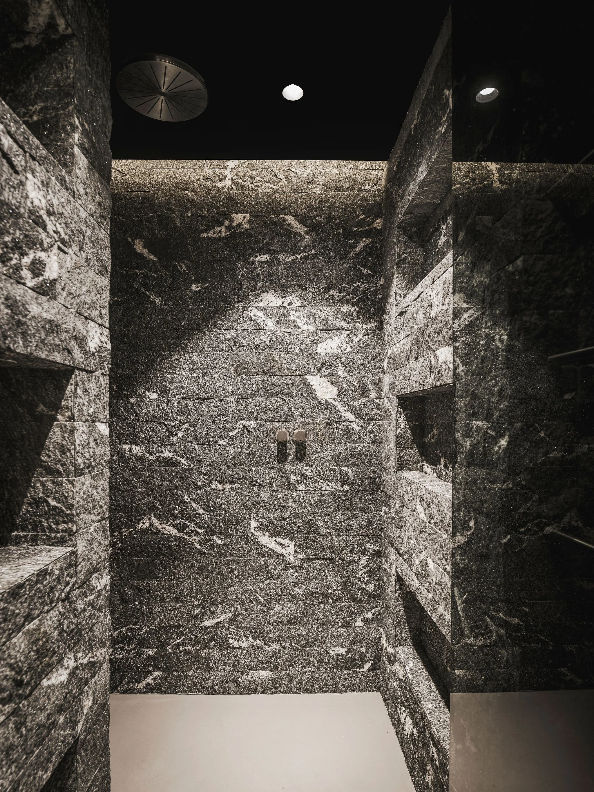 A small, dark room with black marble walls and shelves, two small ceiling lights, and a floor with a light-colored section.