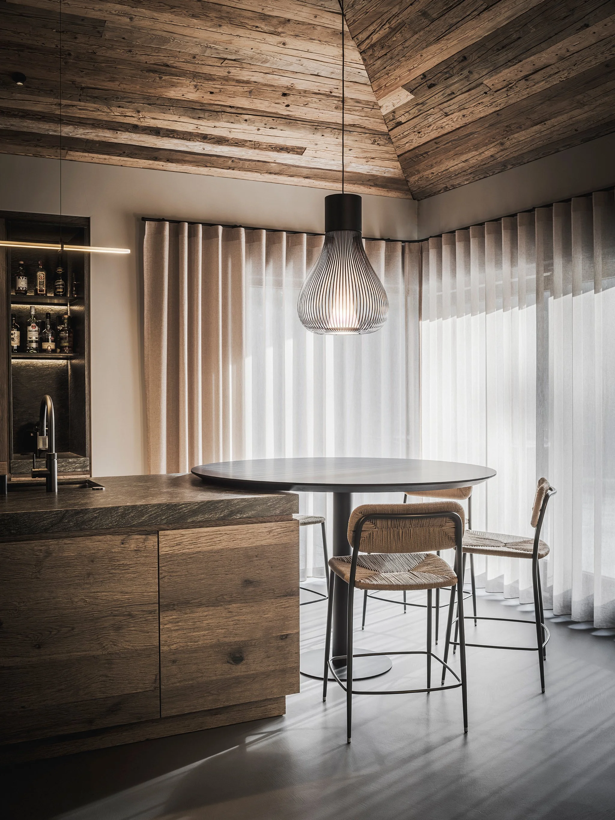 Warm dining area with a round table, two Flexform stools with woven seats, large curtains, and a unique pendant light fixture hanging from a wooden, sloped ceiling.