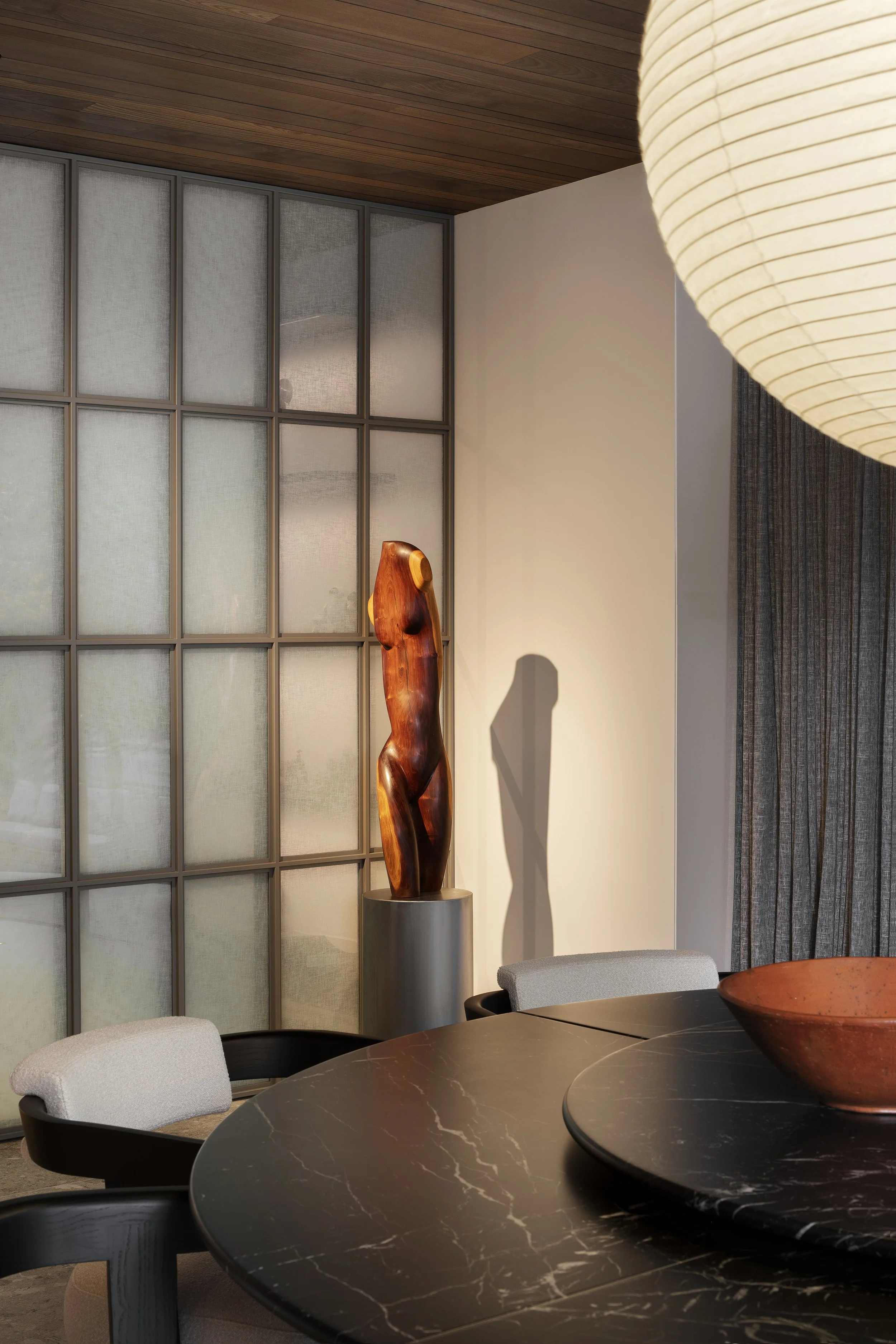 A dining room with a dark marble molteni&C table, light-colored upholstered chairs, a wooden midcentury modern  sculpture on a stand, and a Japanese style window with grid framing. A large paper lantern from Time & style hangs above the table