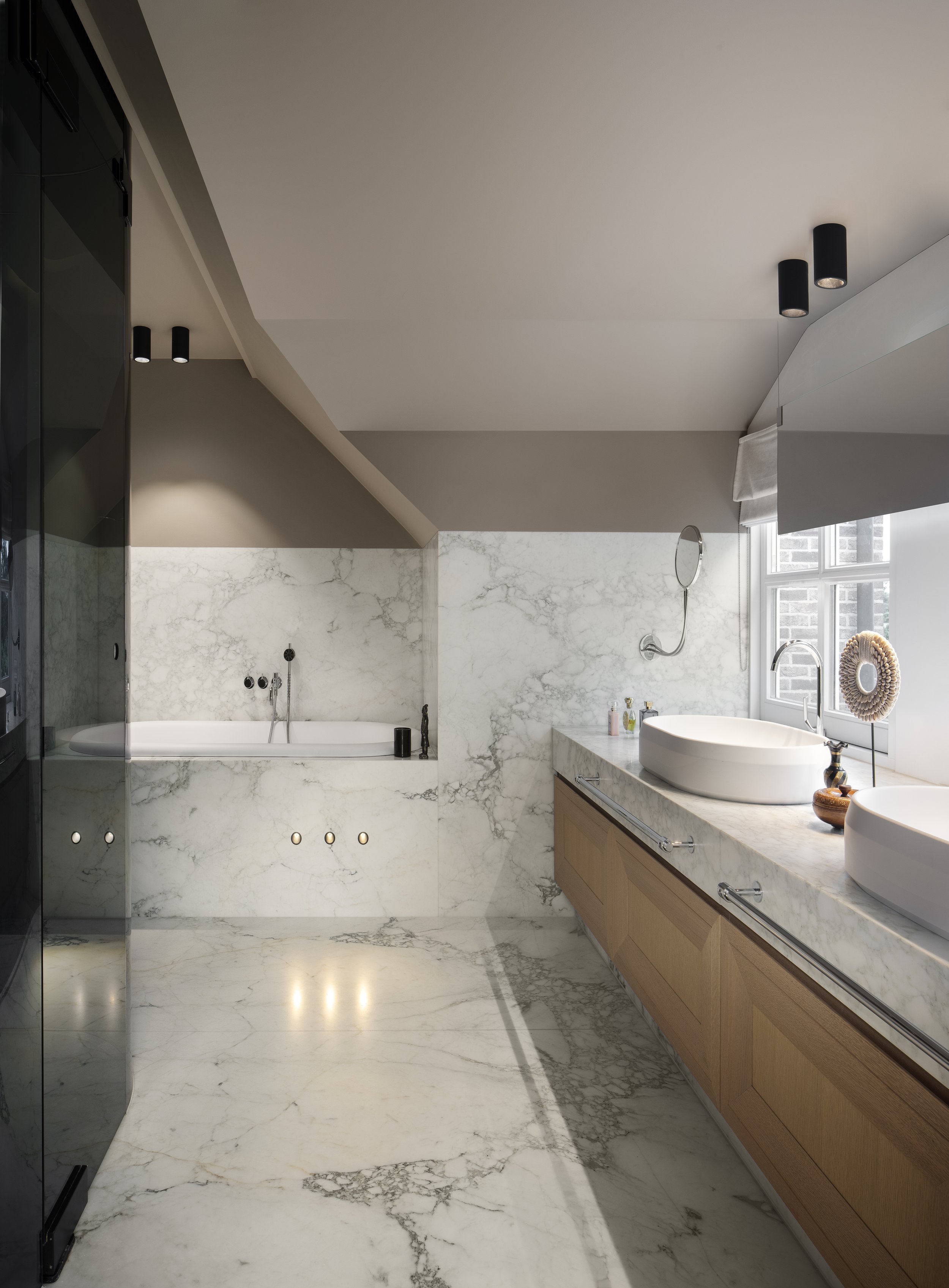 High end design bathroom with marble floors and walls, double sinks on a wooden vanity, a bathtub with marble surround, and a window letting in natural light.