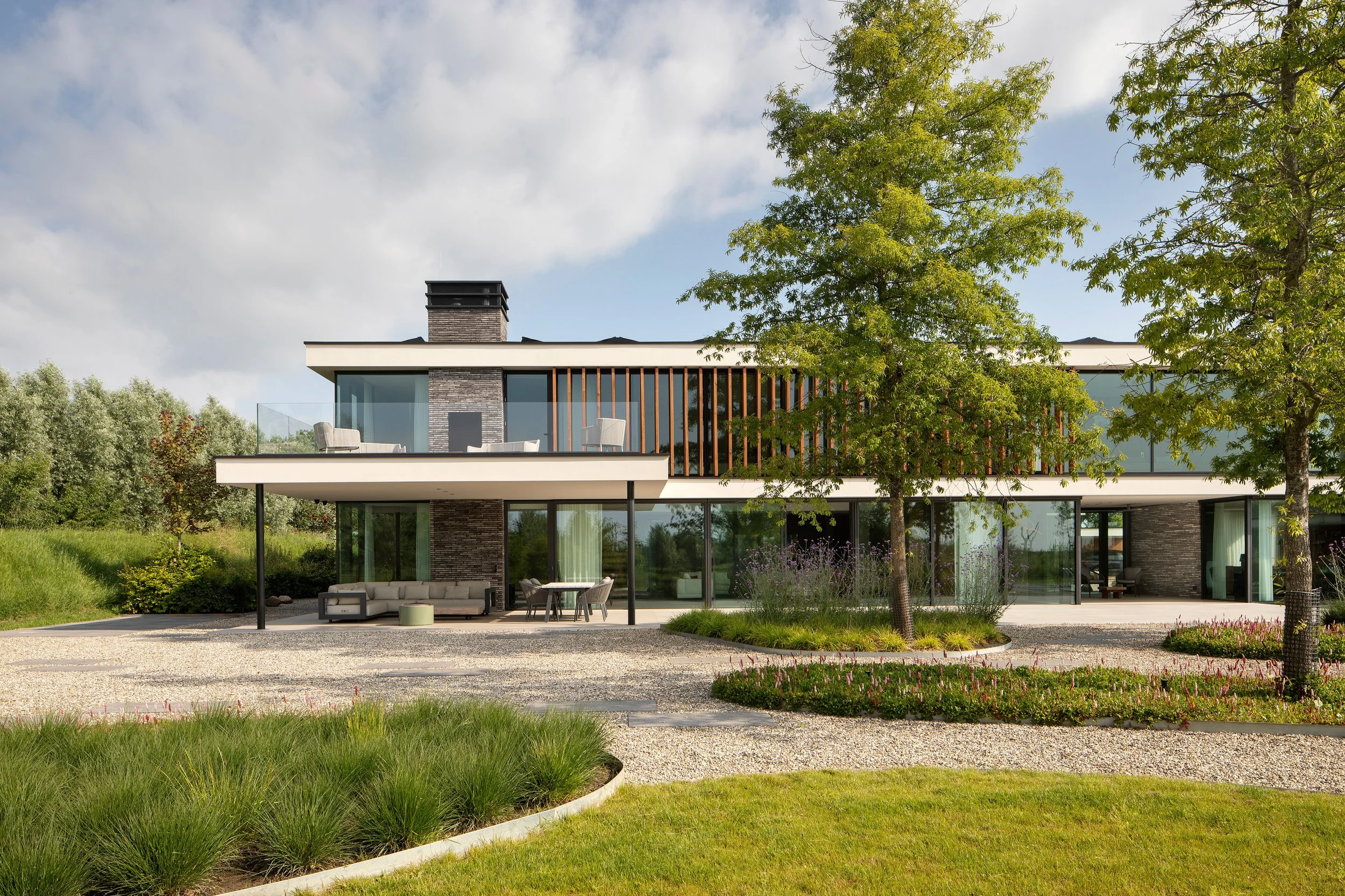 Modern two-story house with large glass windows and balconies, surrounded by landscaped garden with trees, green grass, and pathways under a partly cloudy sky.
