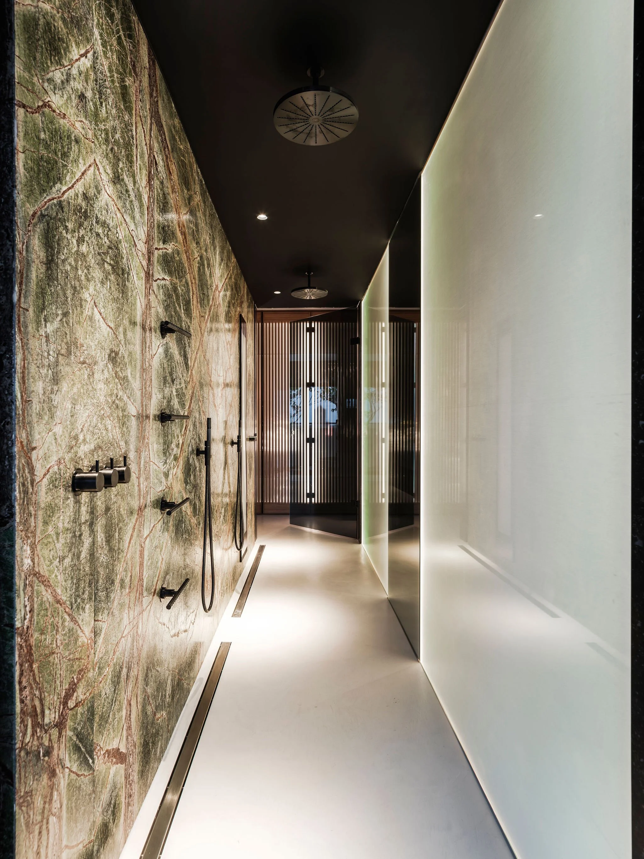 Custom designed luxury shower hallway with marble wall on the left, black ceiling, and frosted glass on the right, leading to wood slatted partitions.