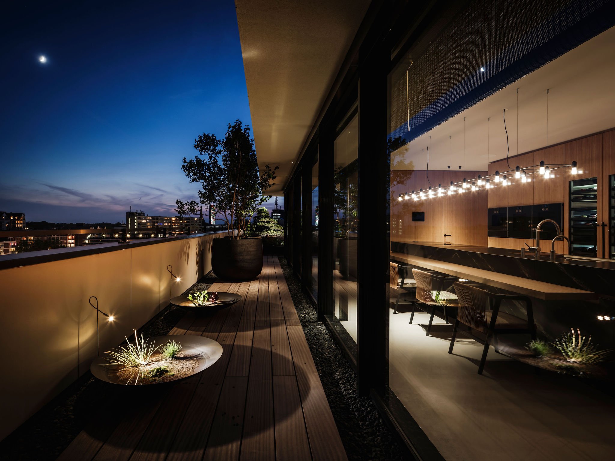 Luxurious rooftop balcony at night with potted plants, wooden flooring, city skyline view, and interior custom designed kitchen with lighting reflections.