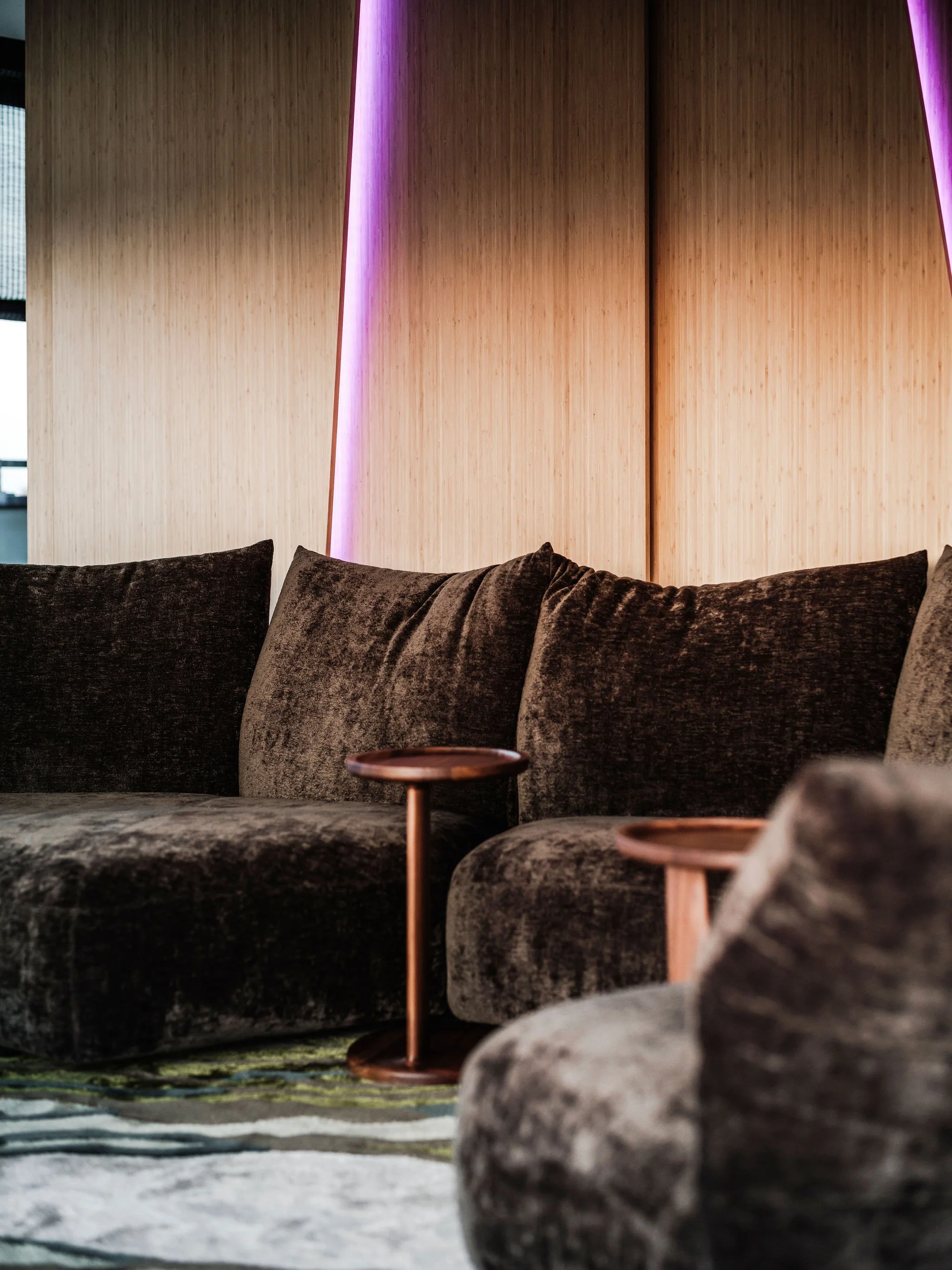 Very high quality living room with plush dark Edra sofas, small round wooden Flexform side tables, and a light bespoke bamboo wood panelled wall with pink accent lighting.