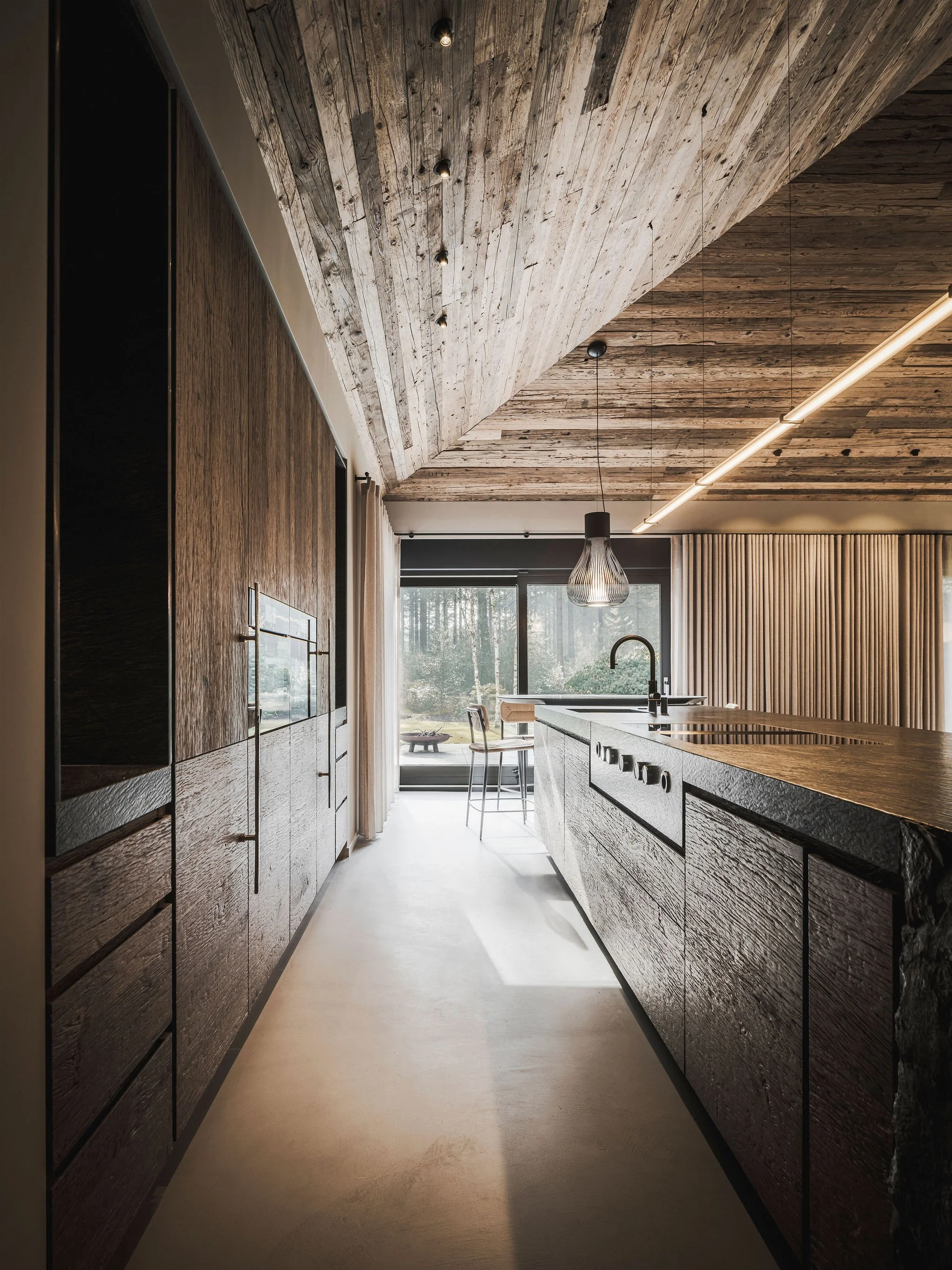 Bespoke Chalet style kitchen with wooden cabinets, a large island with a built-in stove, pendant light, and large window overlooking outdoor scenery.