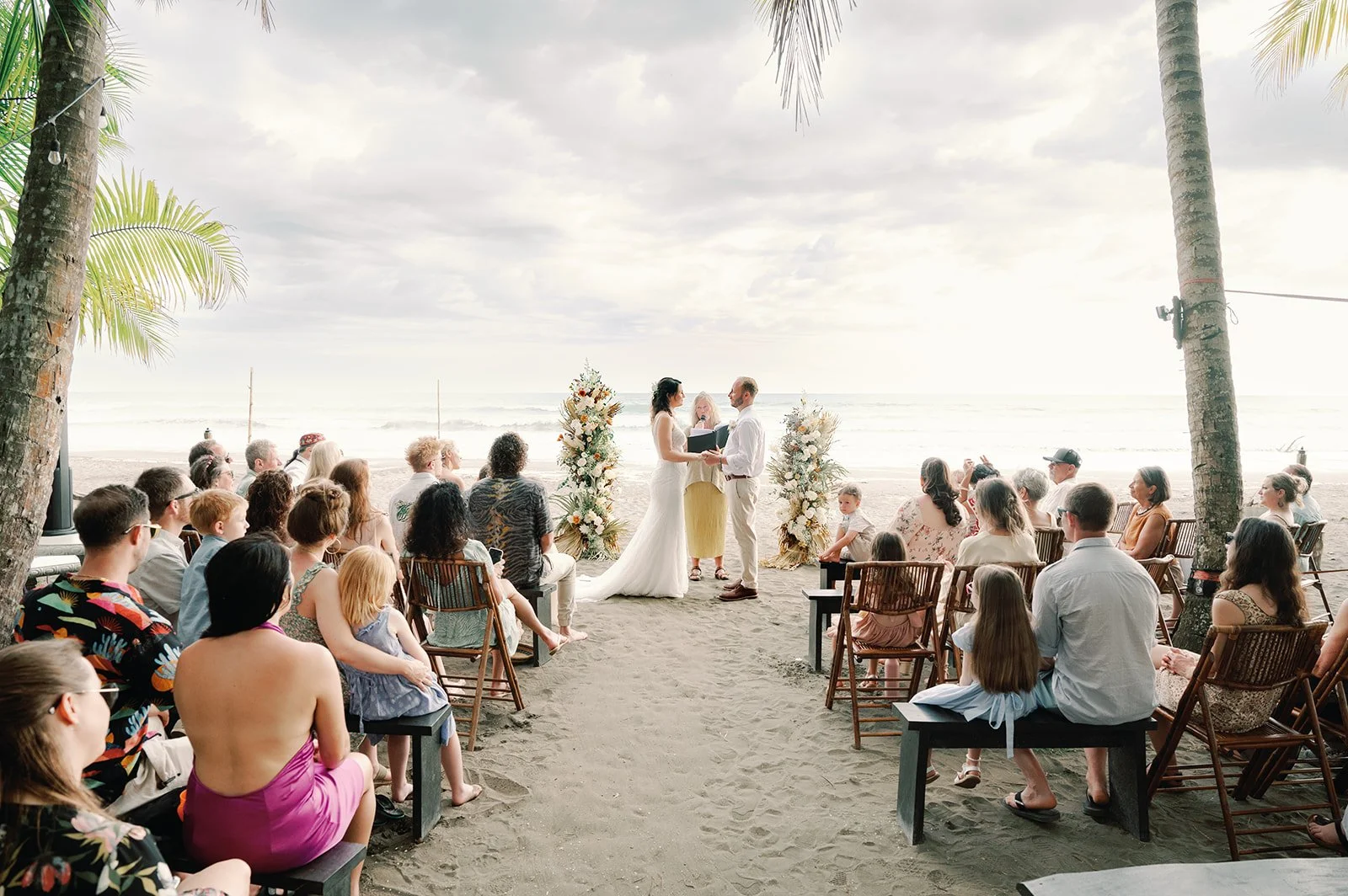 A wedding ceremony taking place on a beach with a bride and groom holding hands, standing in front of an officiant, surrounded by guests seated outdoors. The beach has sand, palm trees, and the ocean in the background, with cloudy skies overhead.