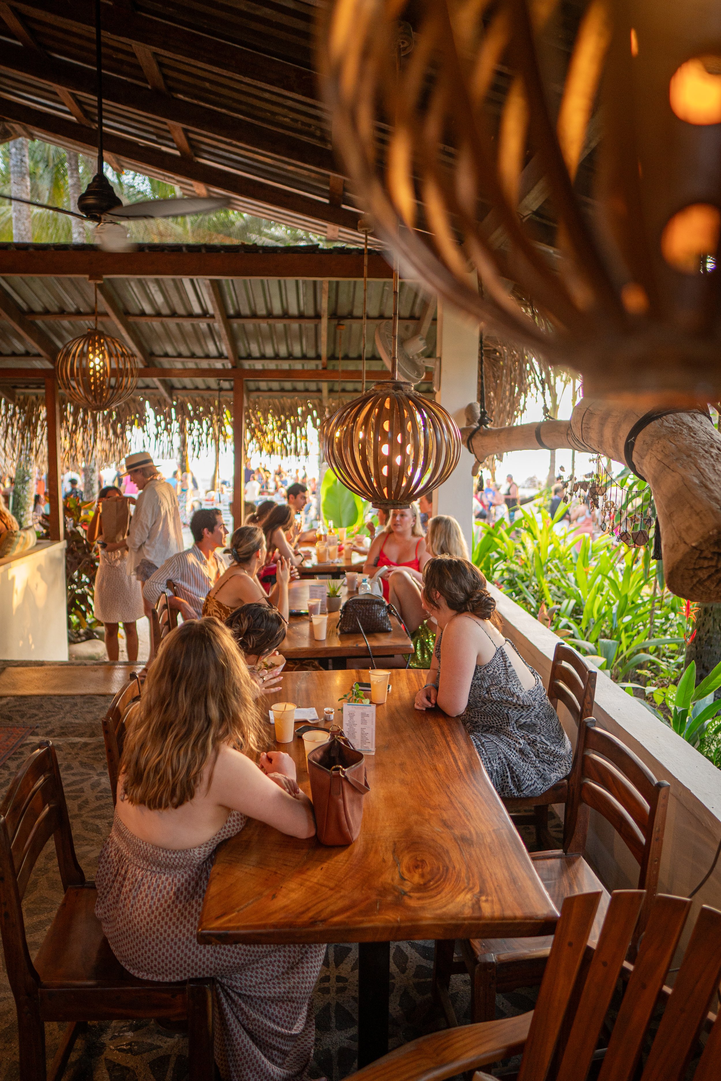 People dining at a restaurant with wooden furniture and tropical decor, hanging woven lamps, lush greenery, and open-air view
