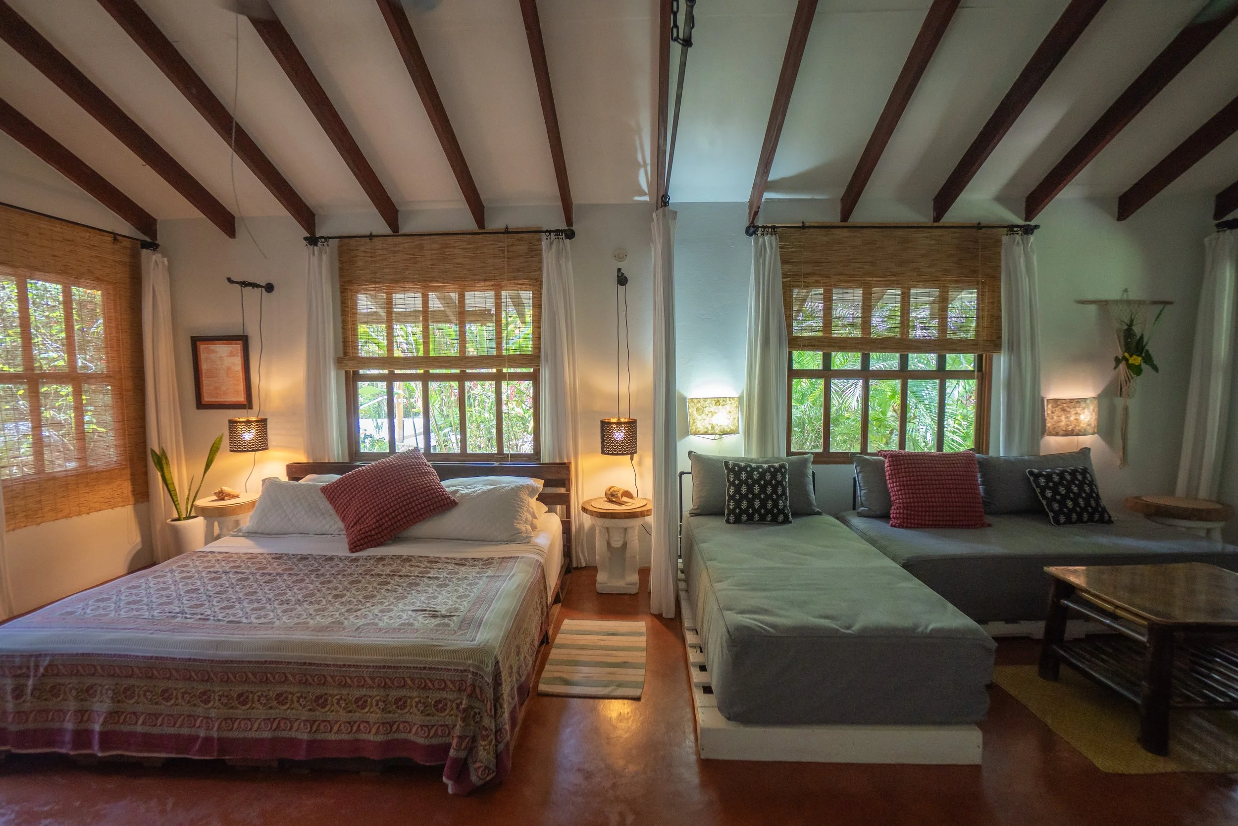 A bedroom with two beds: a larger bed on the left with white bedding and red pillows, and a smaller bed on the right with gray bedding and patterned pillows. The room has large windows with bamboo shades, and there are curtains and decorative plant on the side. Lamps are hung by the beds, and there is wooden roofing with exposed beams.
