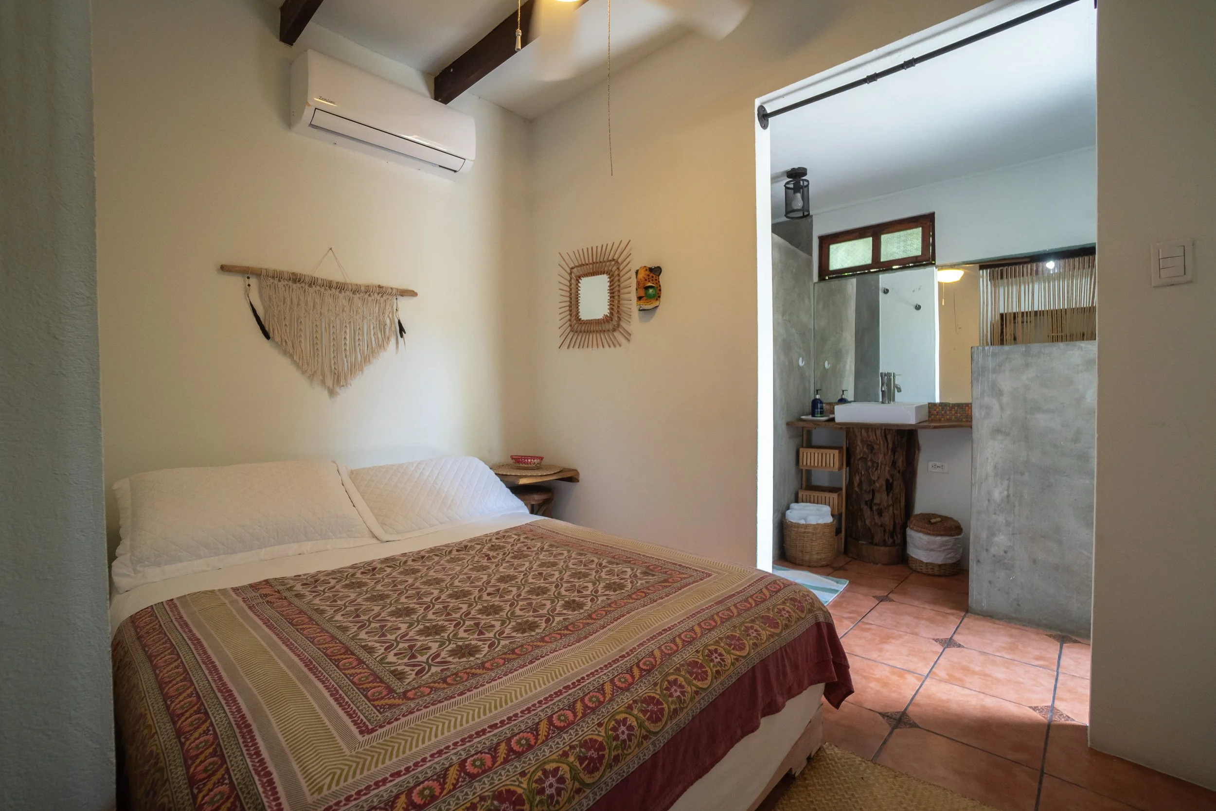 A cozy bedroom with a bed covered in a colorful quilt, wall decorations including a macrame wall hanging and a mirror, an air conditioning unit, and an open bathroom with a sink, wicker baskets, and natural wood accents.
