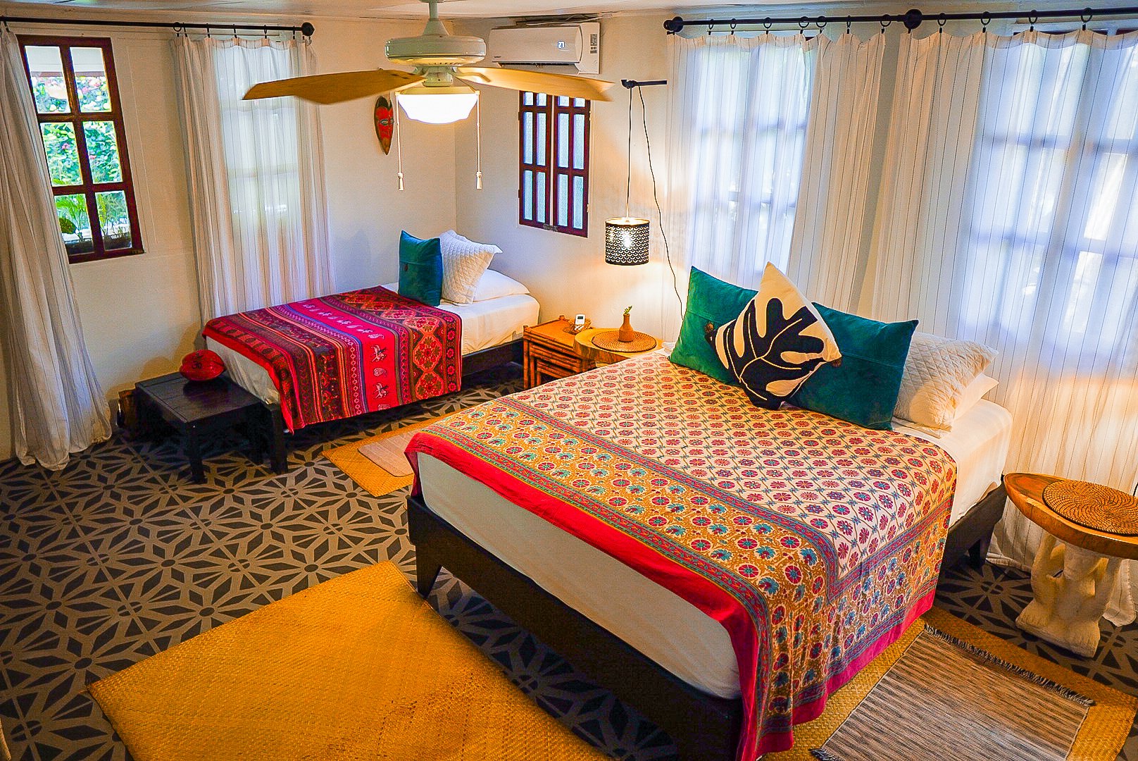 A bedroom with two beds, colorful patterned bedspreads, several pillows, and a small side table with a lamp. There is a ceiling fan, air conditioning unit, and windows with white curtains letting in natural light. The room has a patterned carpet and yellow cushions.