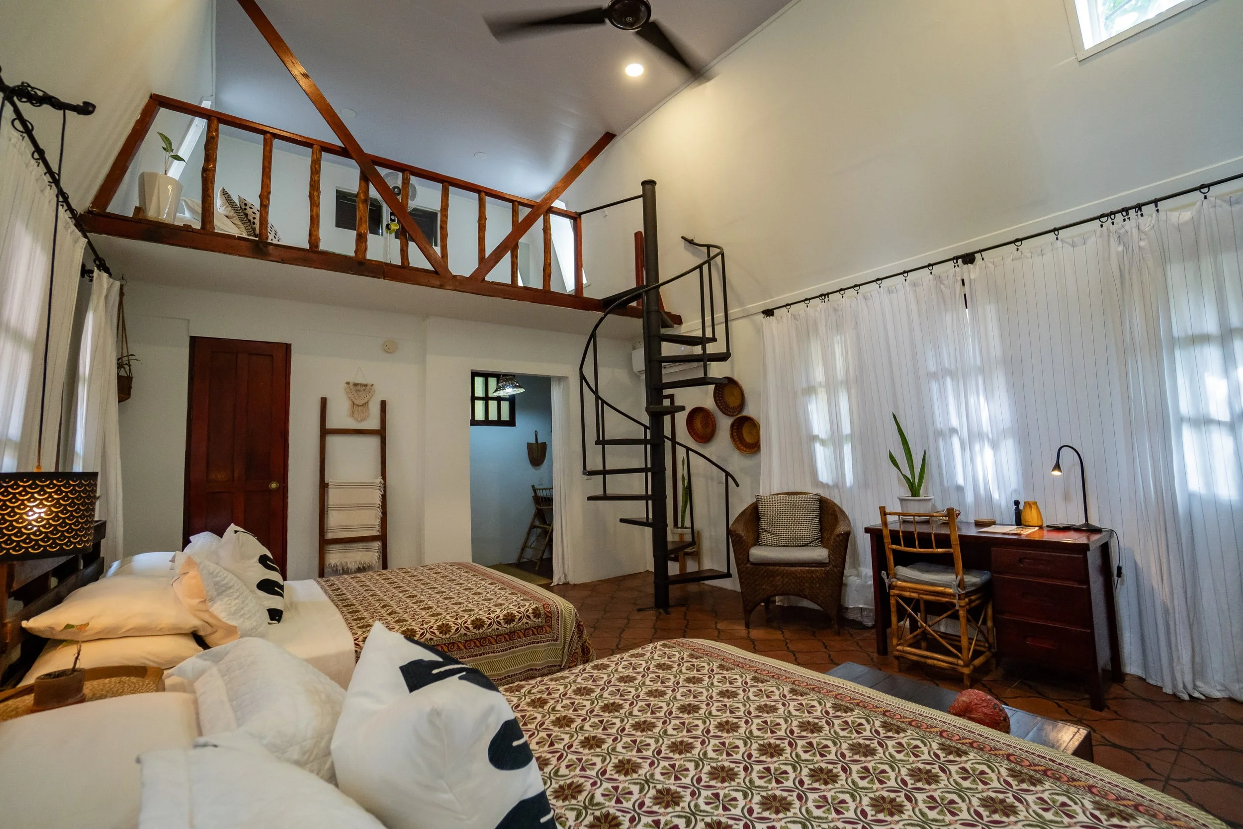 Living room with white walls and large windows with white curtains, a bed with patterned bedding and black and white pillows, a spiral staircase leading to a loft area with wooden railing, a brown wooden door, a brown armchair, a small wooden table, and decorative plates on the wall.