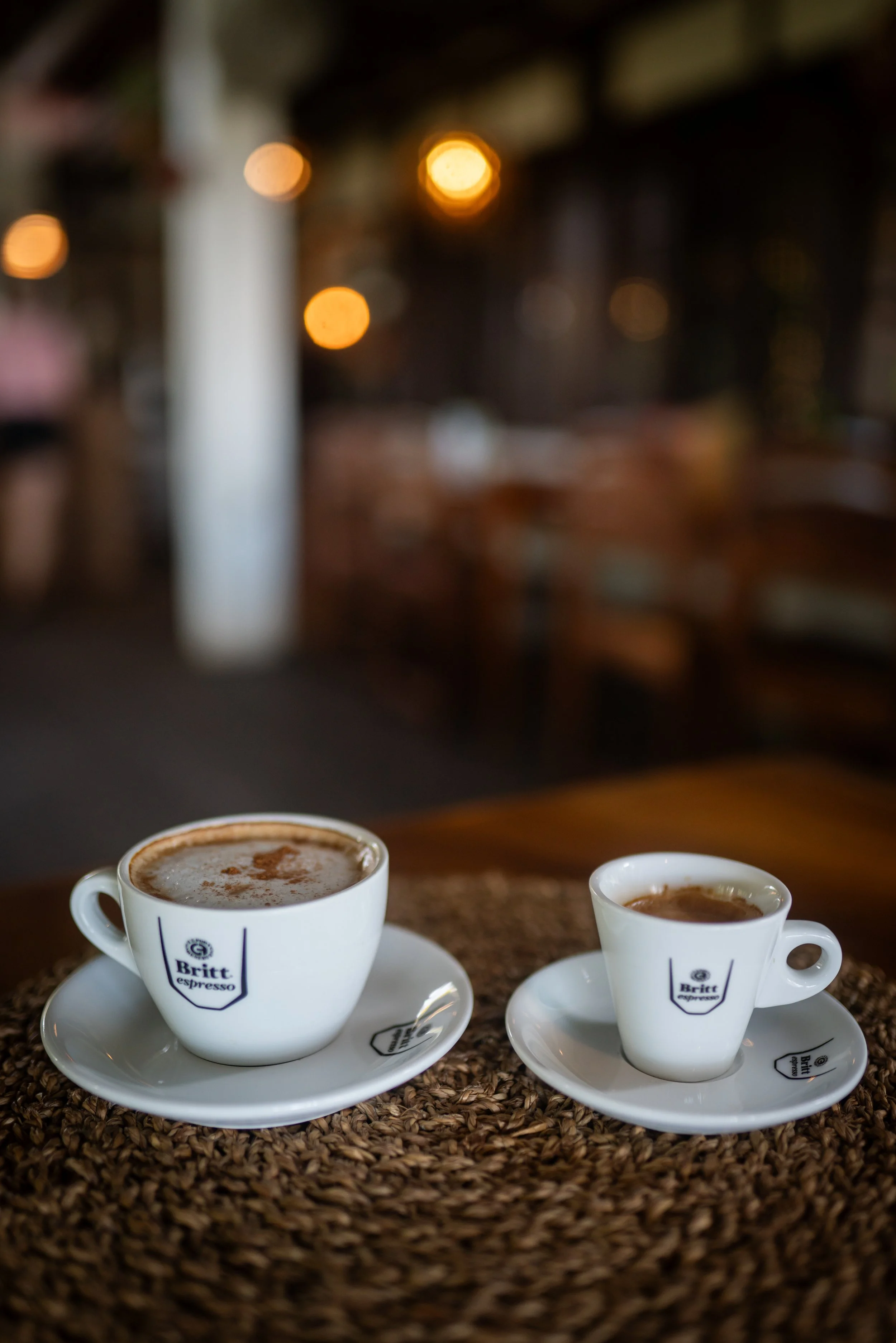 Two white coffee cups with saucers on a table, filled with hot beverages, on a surface covered with coffee beans, with a blurred background of a cozy cafe interior with warm lights.