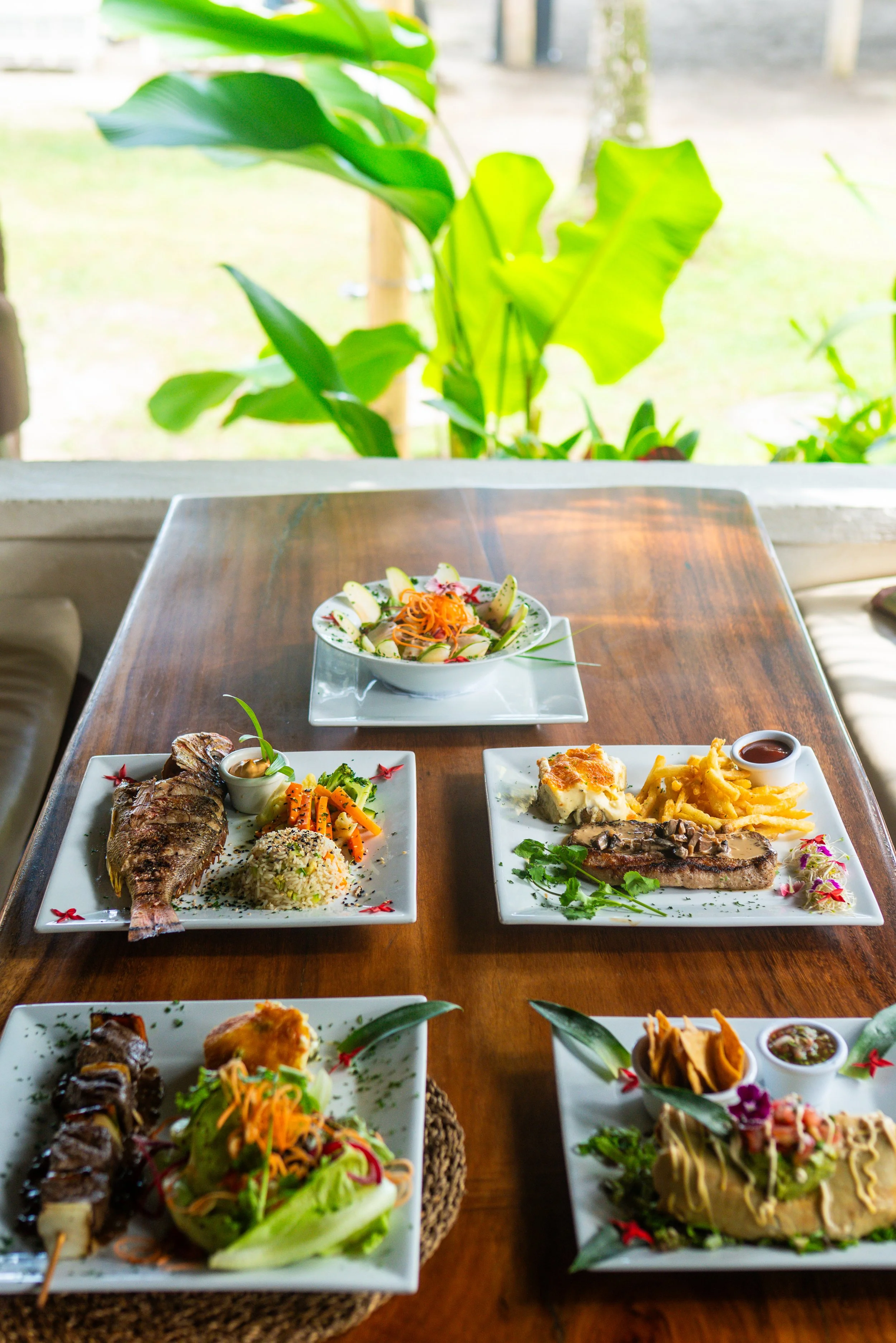A variety of plated Asian dishes arranged on a wooden table with a lush green outdoor background.