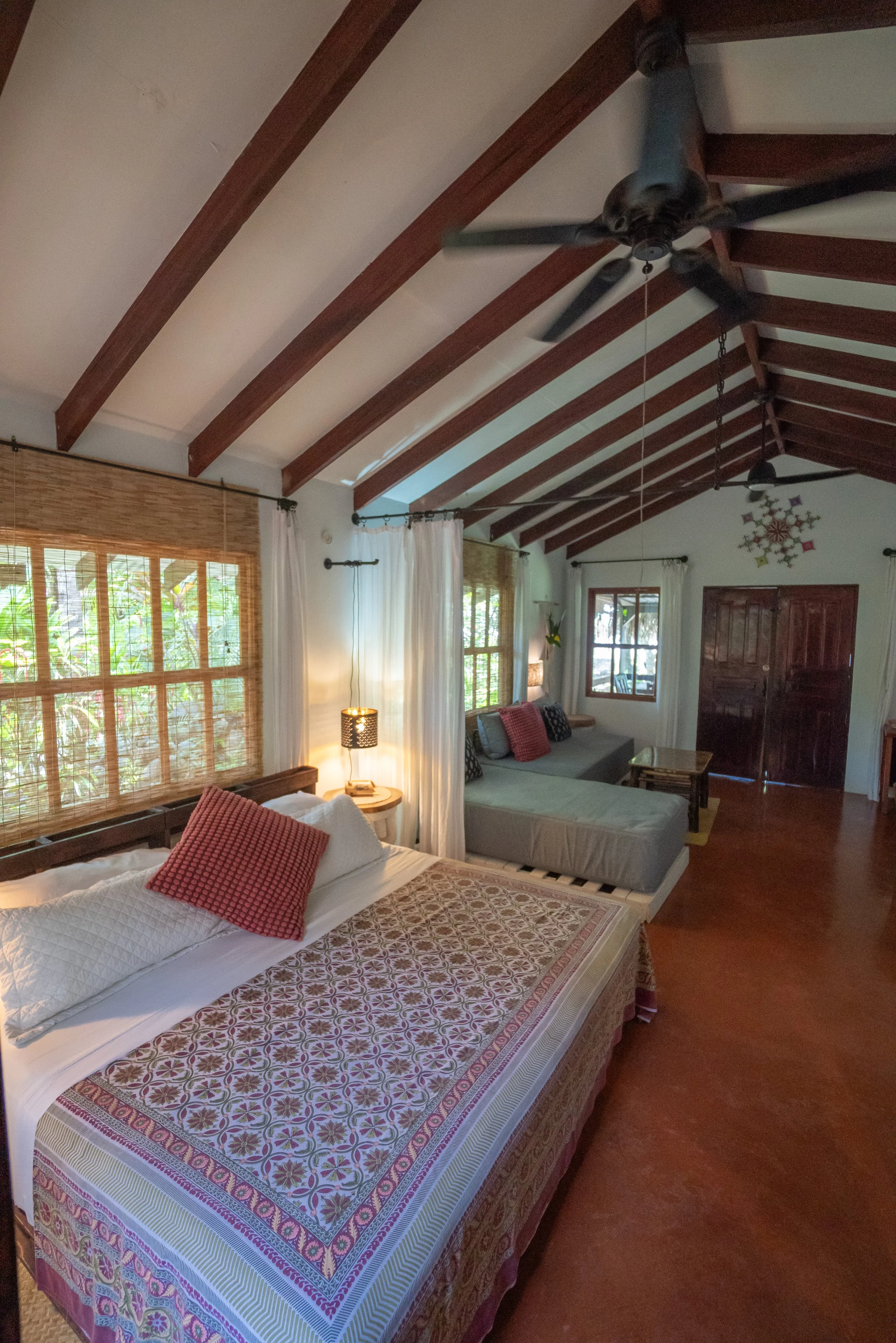 A bedroom with a bed covered in a colorful patterned bedspread, multiple pillows, and a window with bamboo blinds. There is a gray couch with cushions, a side table with a lamp, and wooden beams on the ceiling. The room has white curtains and wooden furniture.