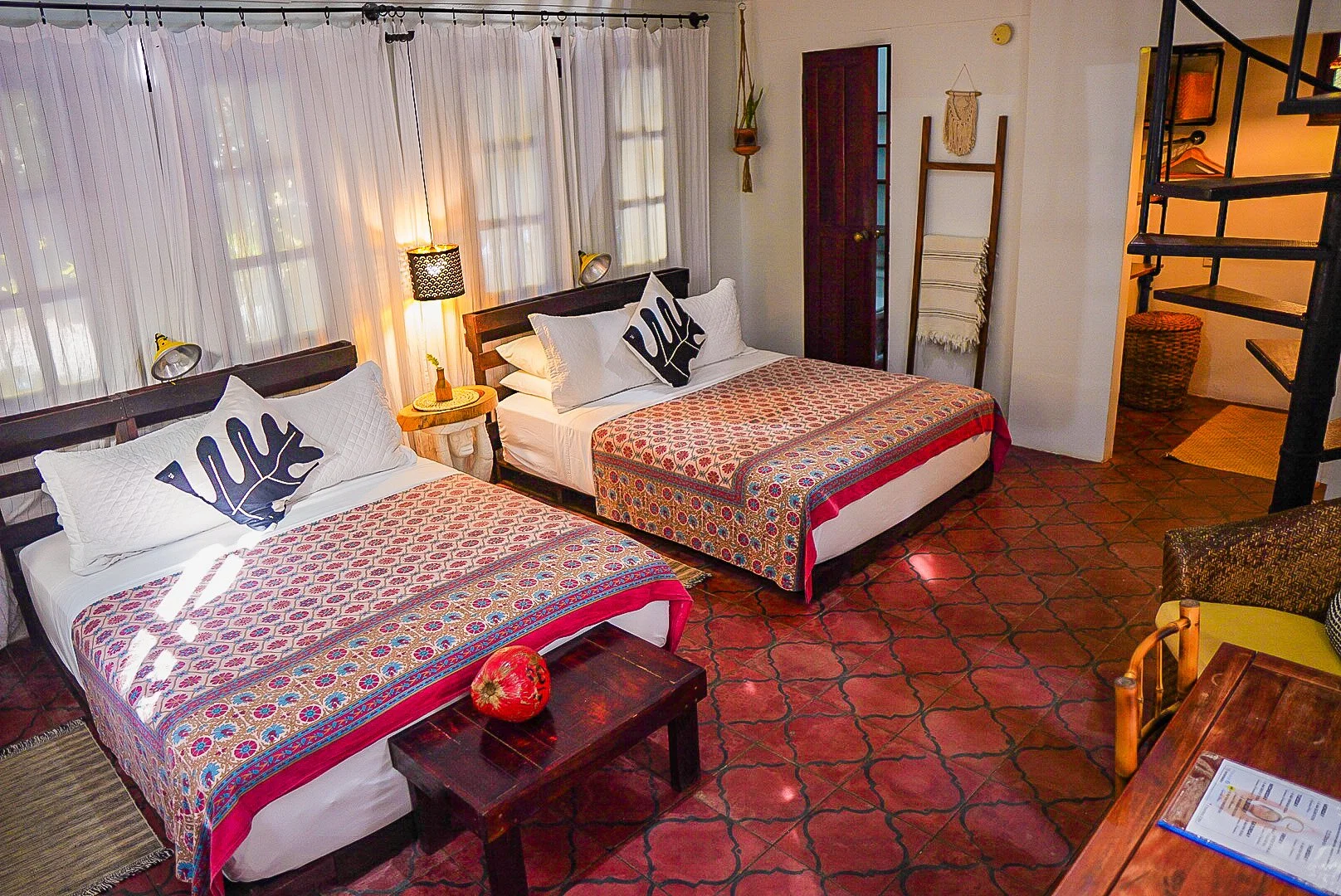 A cozy hotel room with two twin beds featuring colorful patterned bedspreads, decorative pillows, and wooden headboards. There are bedside lamps, a small wooden side table, and a window with sheer white curtains. The room has terracotta tile flooring, a wooden door, wall hangings, and an open staircase leading upstairs.
