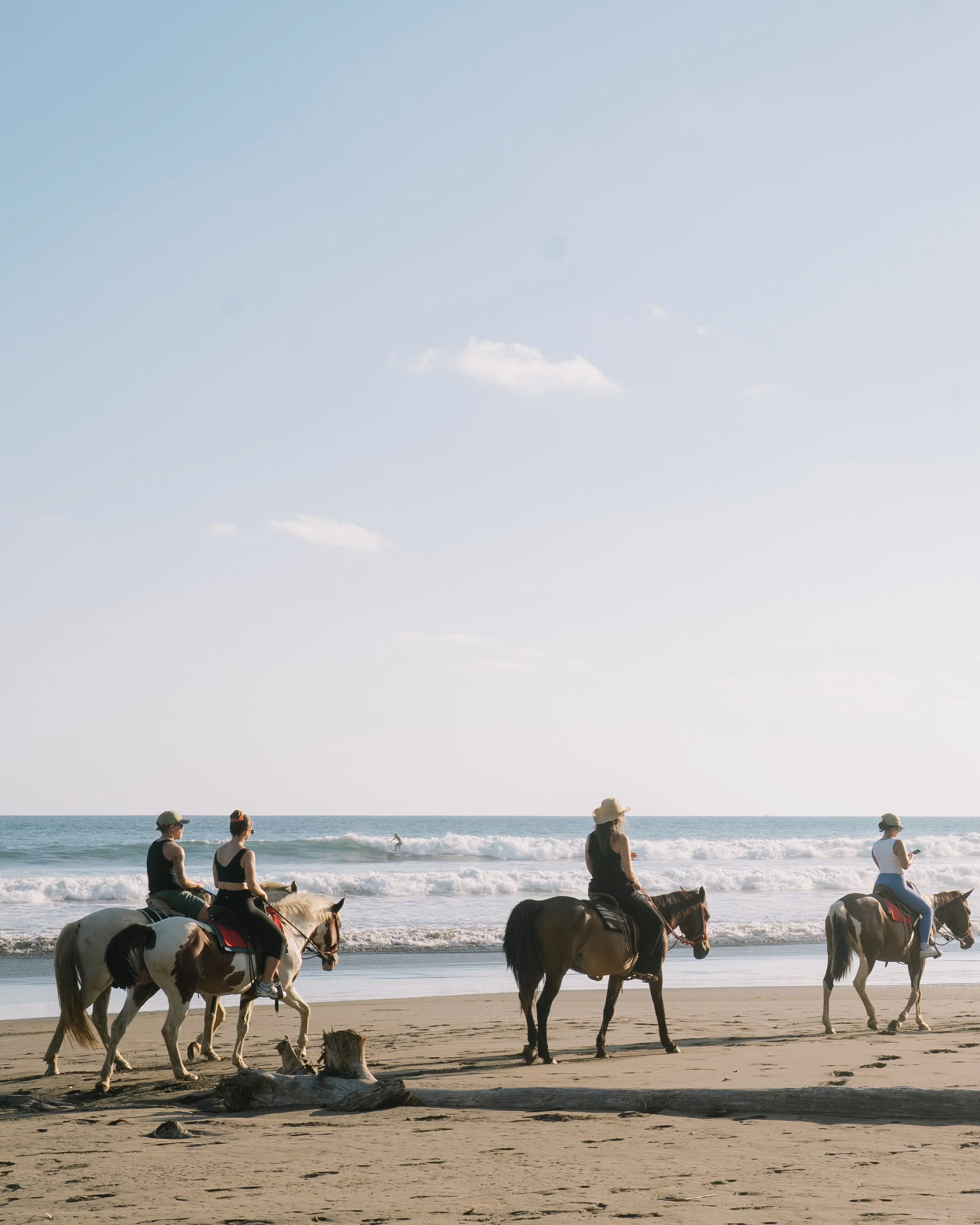 People riding horses along the beach near the shoreline with the ocean in the background and a partly cloudy sky overhead.