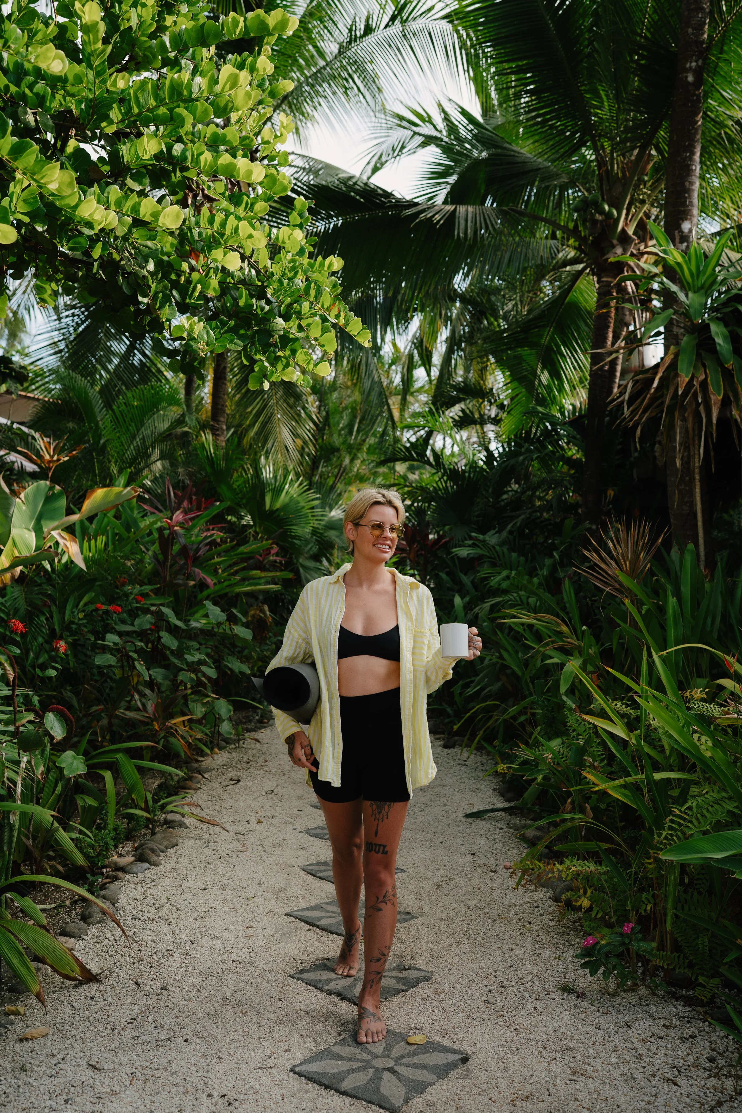 A woman walking barefoot on a stone path through lush tropical greenery, holding a white mug in one hand and a black yoga mat in the other, wearing a yellow button-up shirt over a black bra and black shorts, with tattoos on her thighs.