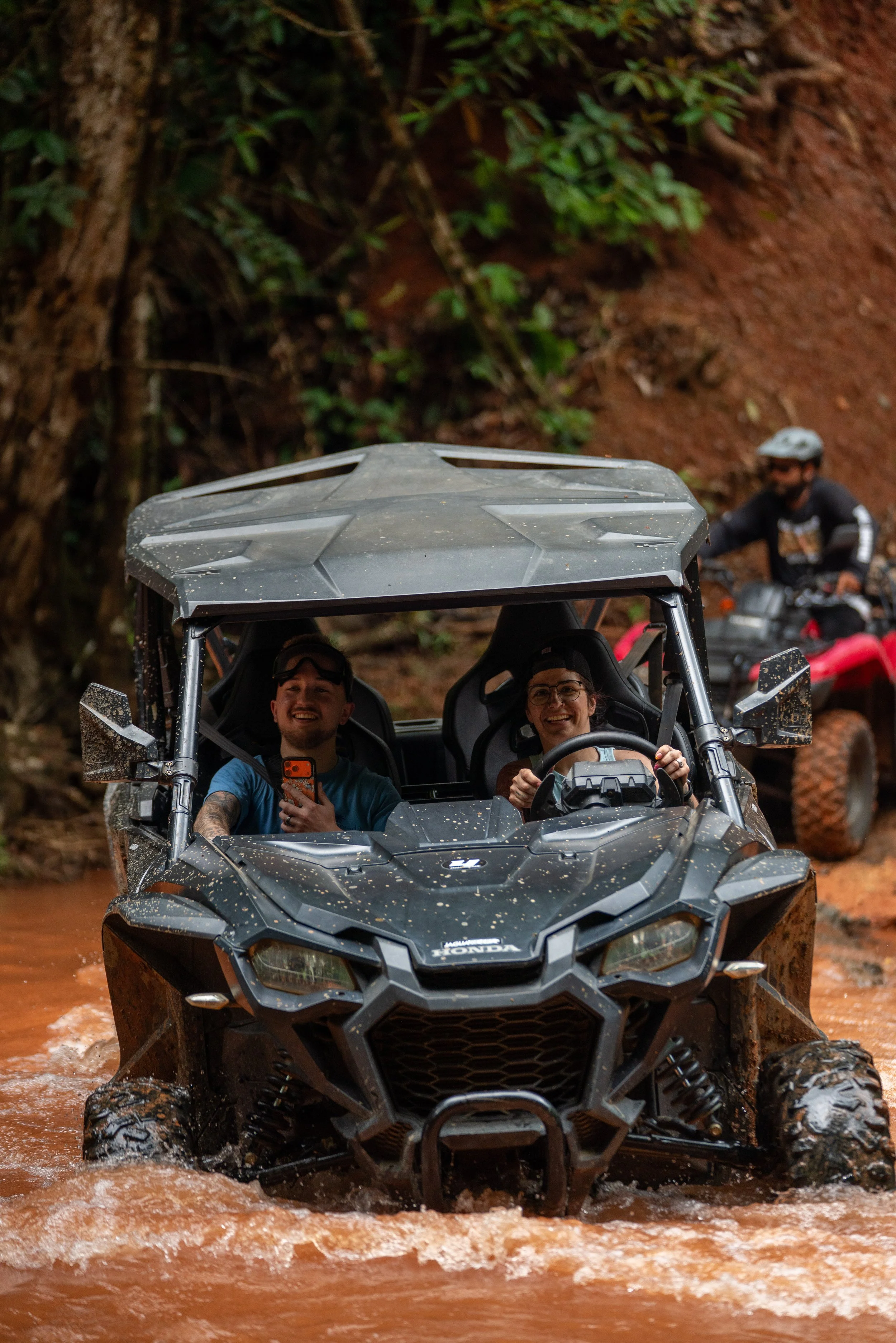 A group of people riding a black Honda off-road utility vehicle through a muddy water crossing in a forest setting, with one person in the front smiling and holding a phone, and another behind wearing a helmet.