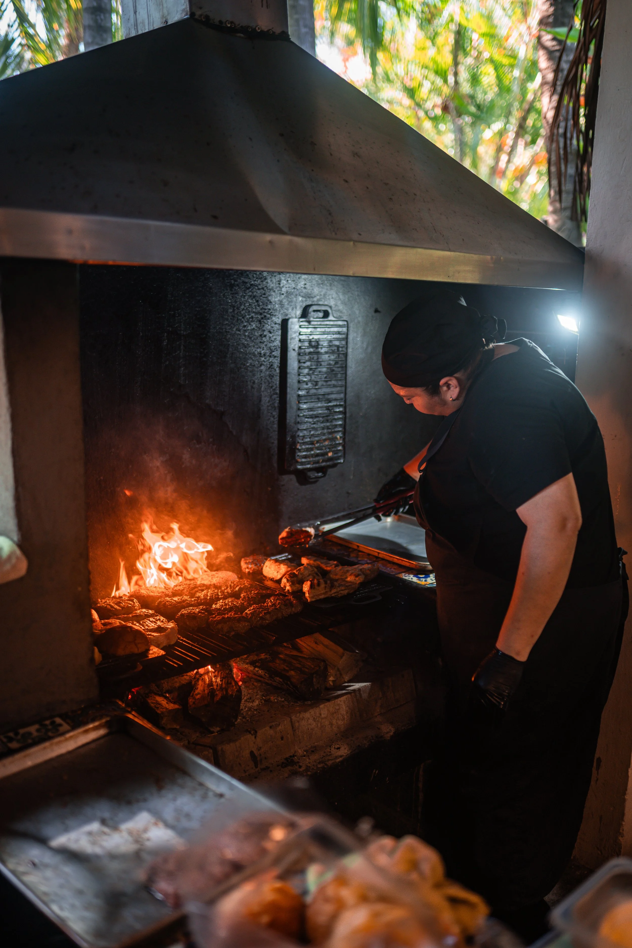 A person cooking on a grill with food over an open flame. The person wears a black outfit and hairnet, seen from the side, inside a kitchen with an outdoor view of green trees.
