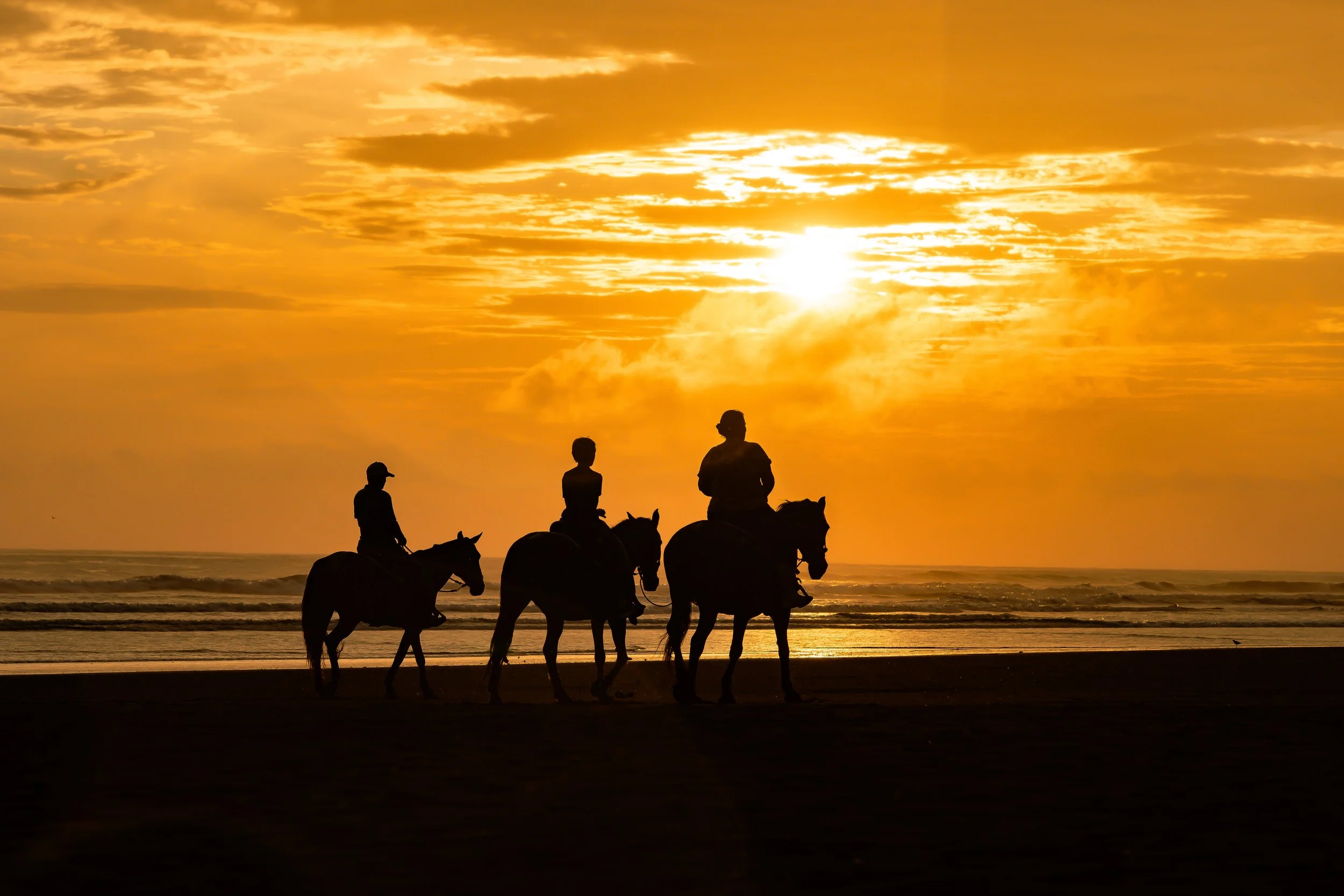 Silhouettes of five people riding horses along a beach during sunset with a bright sun and partly cloudy sky.