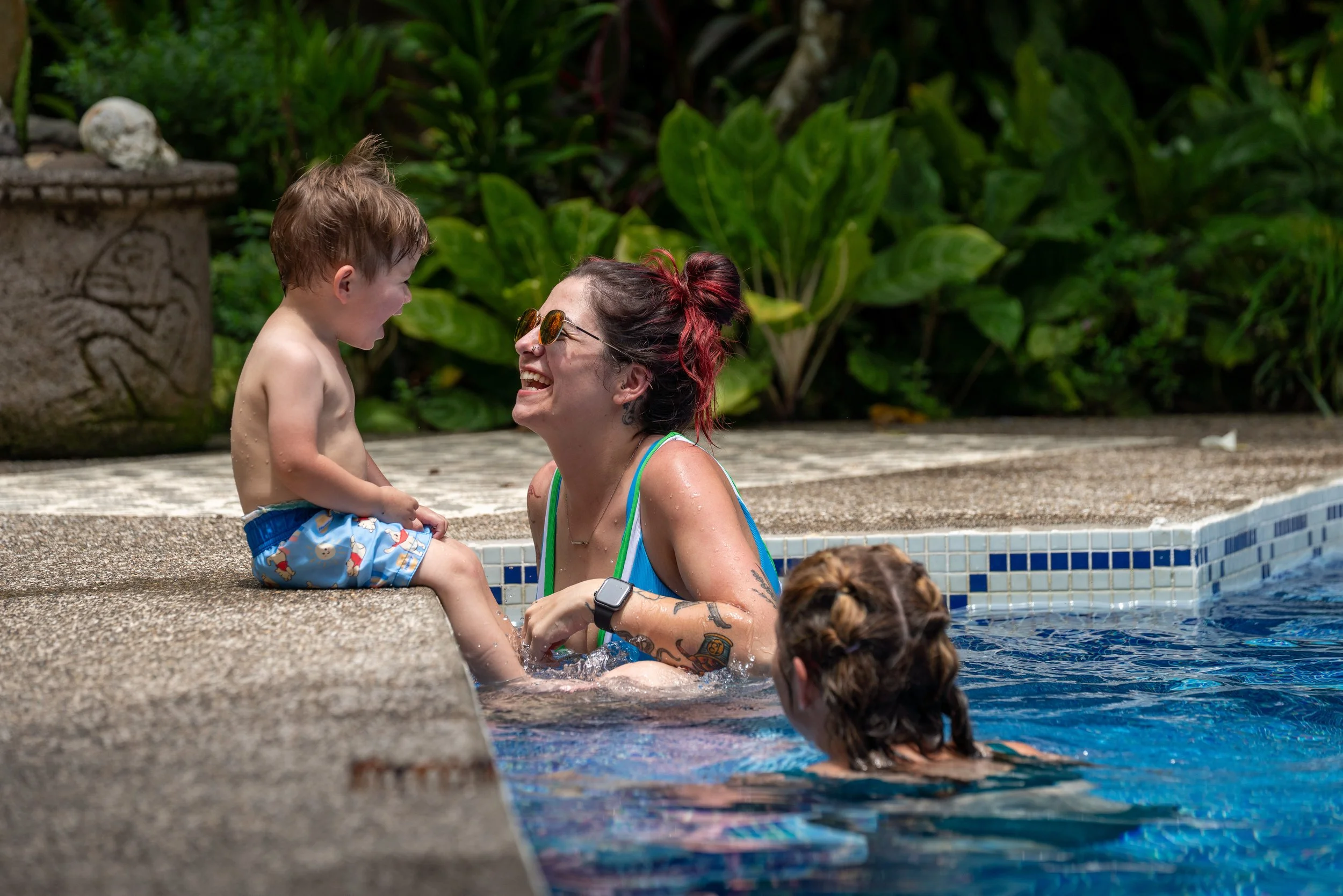 A woman with red hair and sunglasses smiling in a swimming pool, holding a young boy with wet hair and colorful swim trunks, on the pool edge with lush green plants in the background.