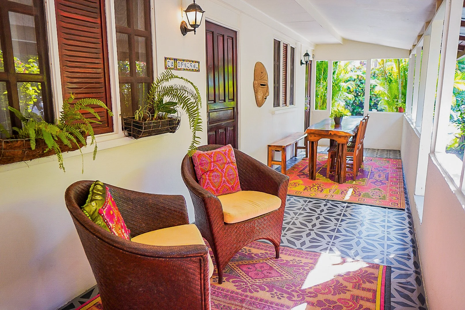 Enclosed porch with wicker chairs, colorful cushions, a wooden table with chairs, potted plants, patterned rugs, and decorative wall hangings, overlooking lush greenery.