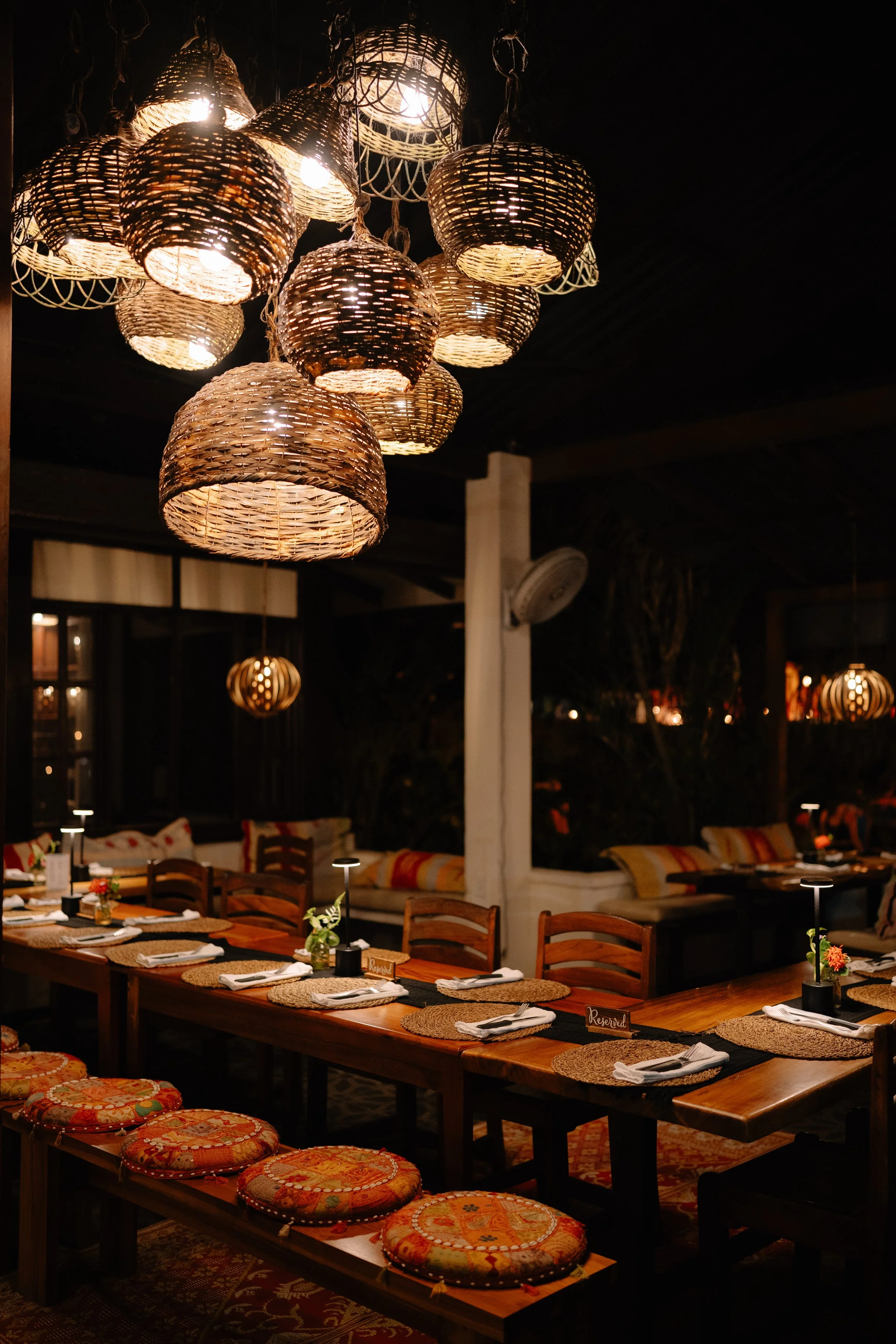 Interior of a restaurant with hanging woven rattan lamps, a long wooden table set for dining, small flower arrangements, and cushioned stools with embroidered fabric seats in a warm, cozy ambiance.