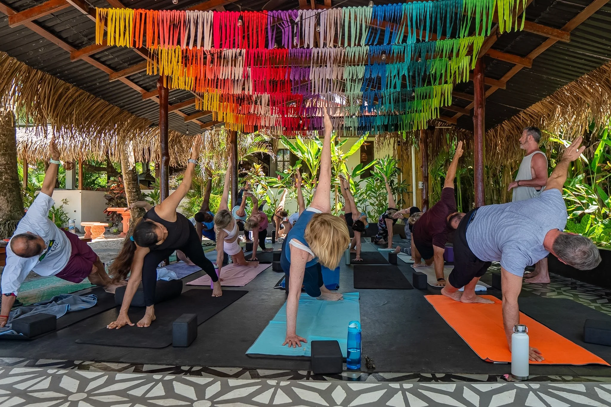 People participating in a yoga class outdoors under a thatched roof, with colorful paper decorations hanging from the ceiling.
