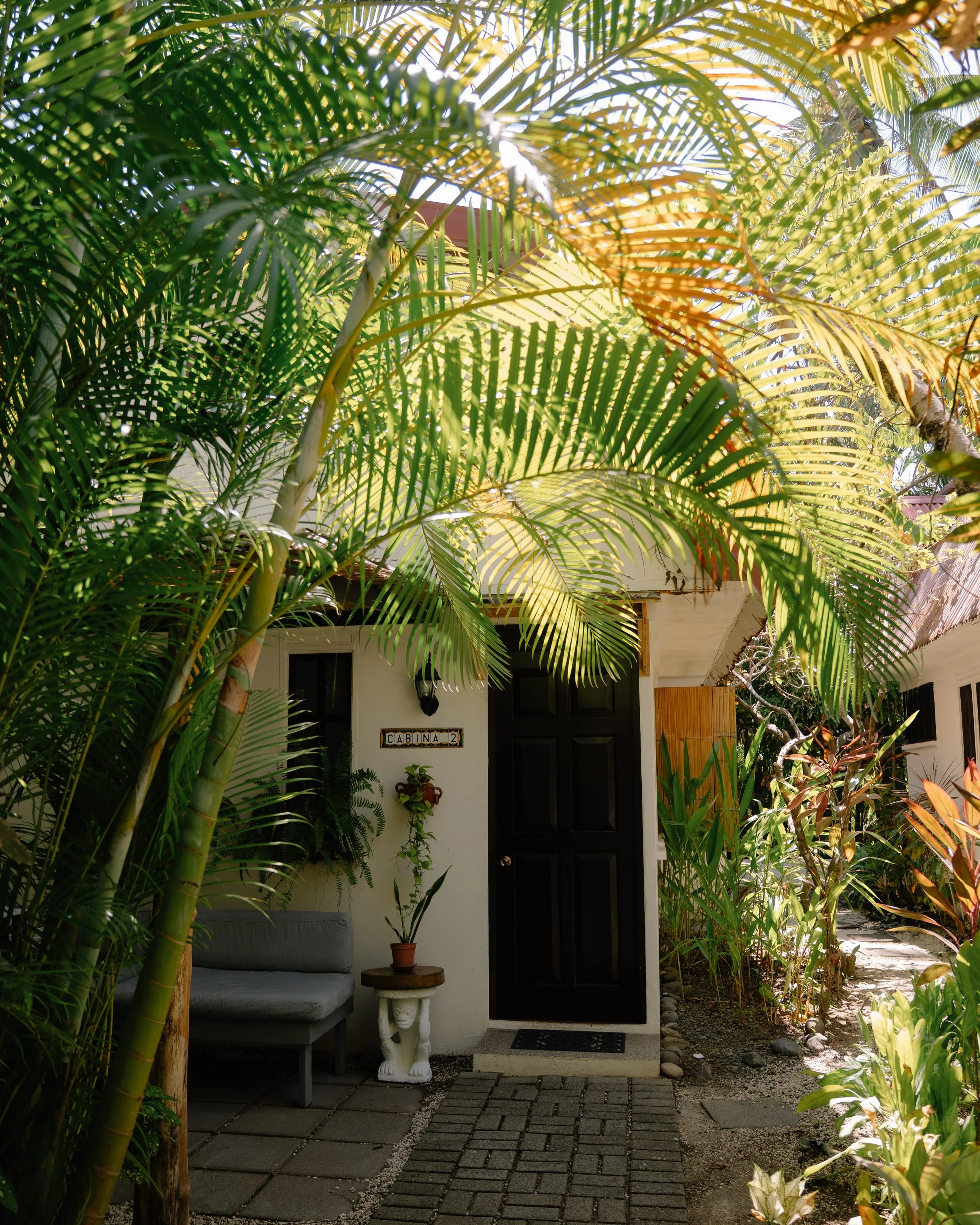 Front door of a house with the sign 'CABINA 2', surrounded by lush green plants and outdoor furniture, including a gray loveseat, a small round table with a potted plant, and a pathway made of brick pavers.