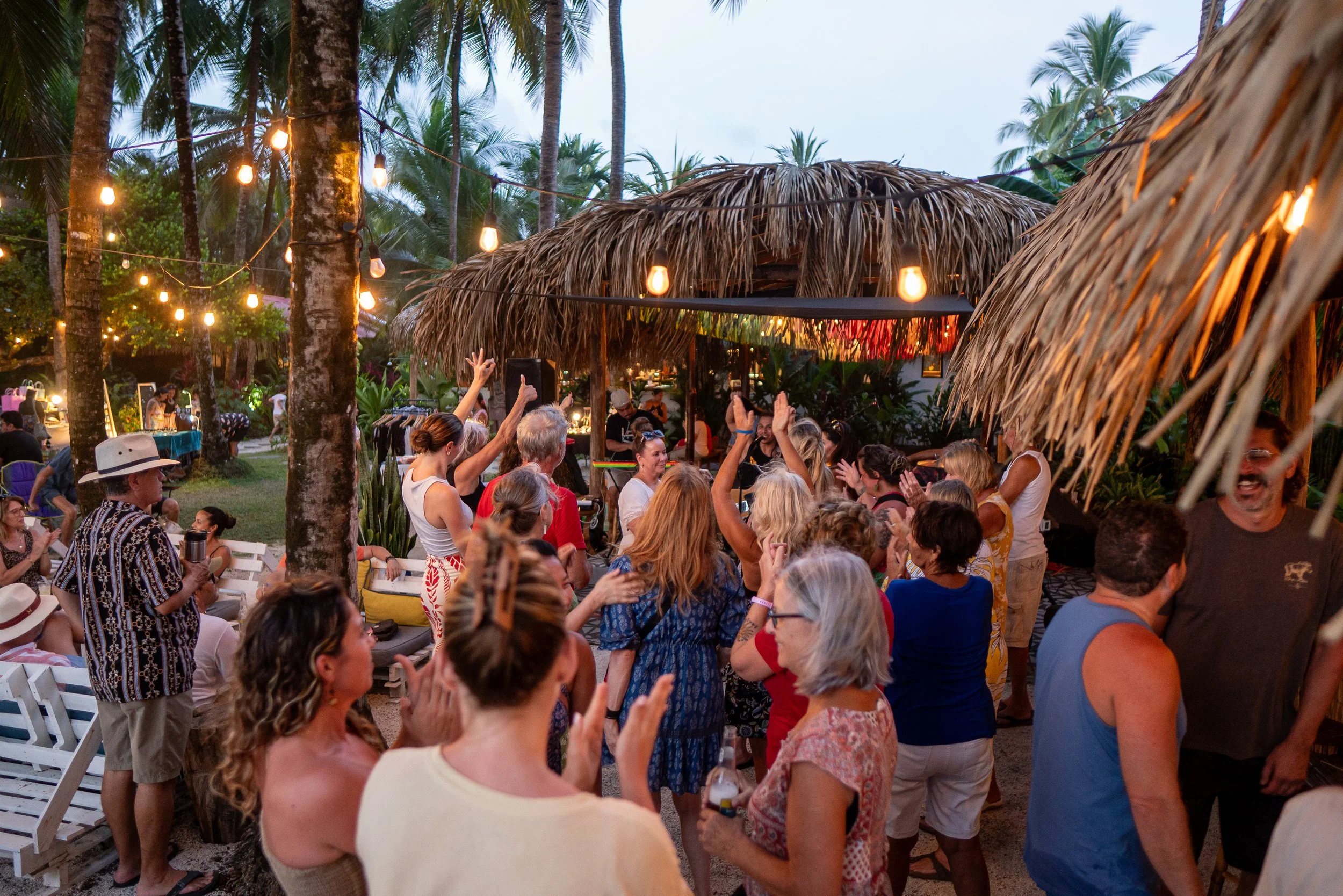 A lively outdoor gathering of people dancing and enjoying music at a tropical beachside venue decorated with string lights and thatched roof structures, surrounded by palm trees.