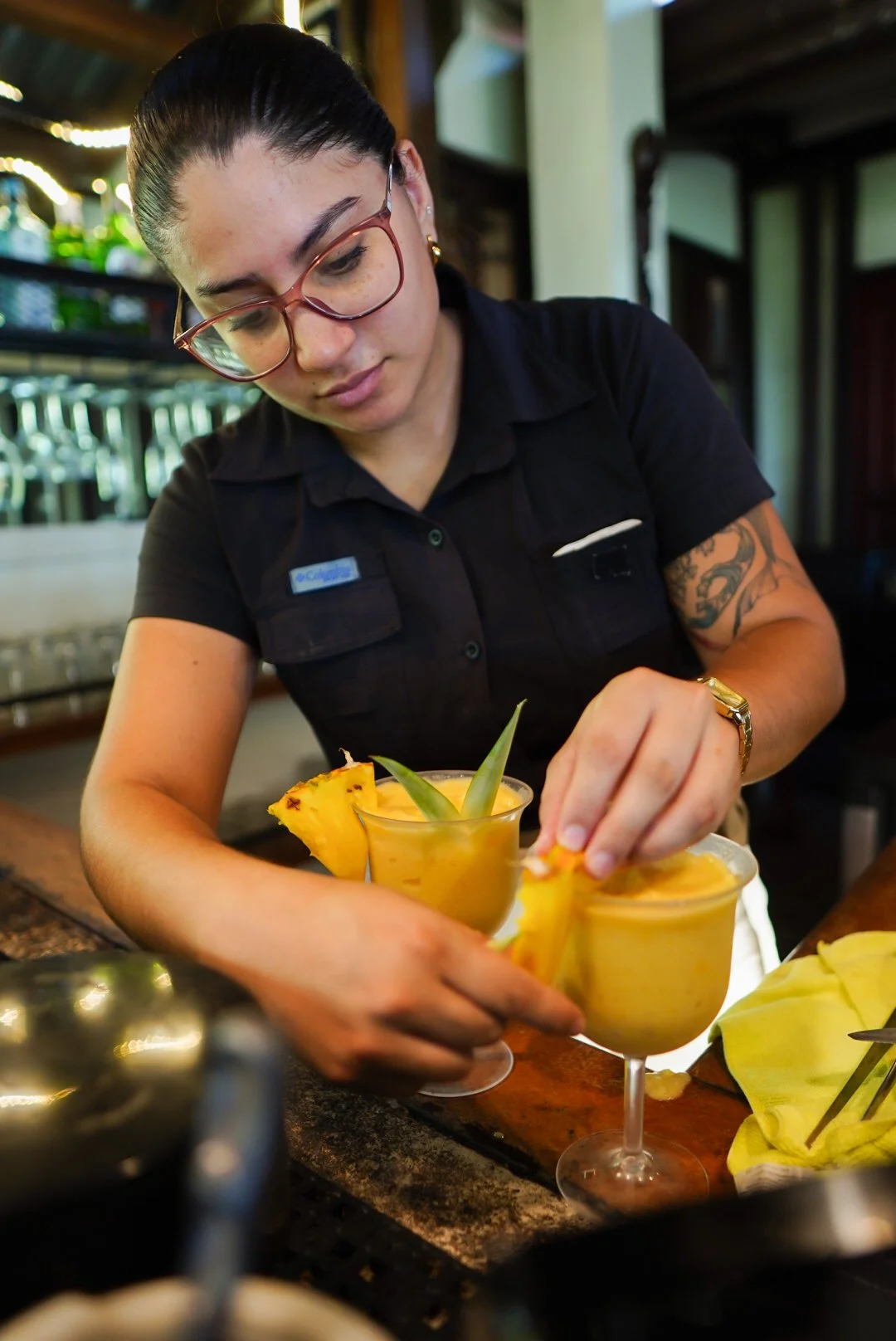 A woman with glasses and a tattoo on her arm is preparing tropical fruit drinks with pineapple garnish in a bar or restaurant.