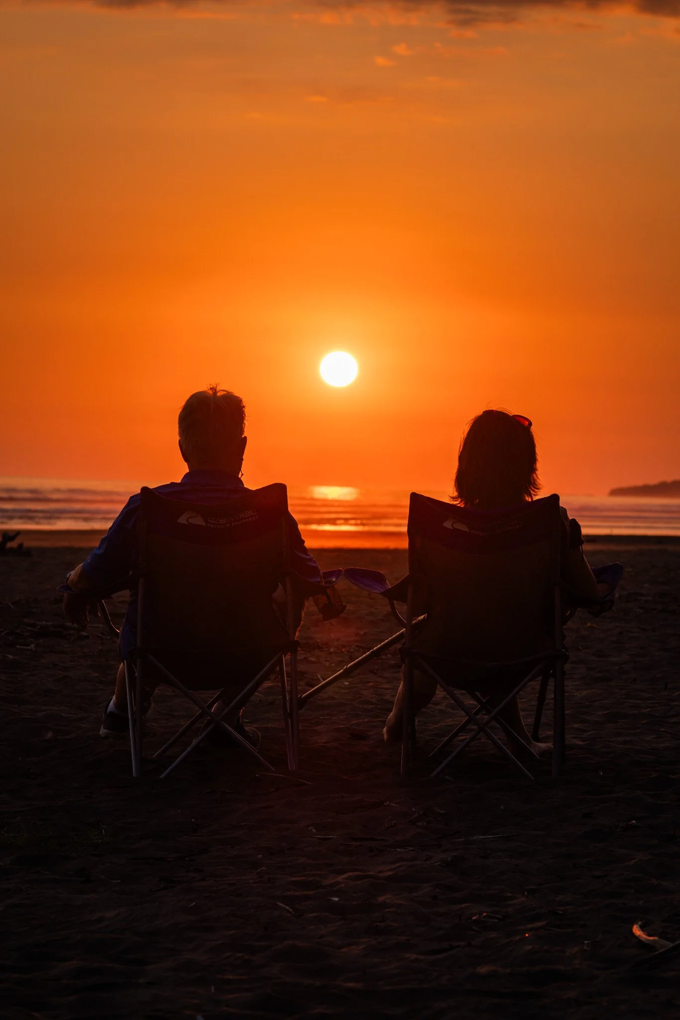 Two people sitting in chairs on the beach during sunset, facing the ocean with the sun low on the horizon, and enjoying the scenic view.