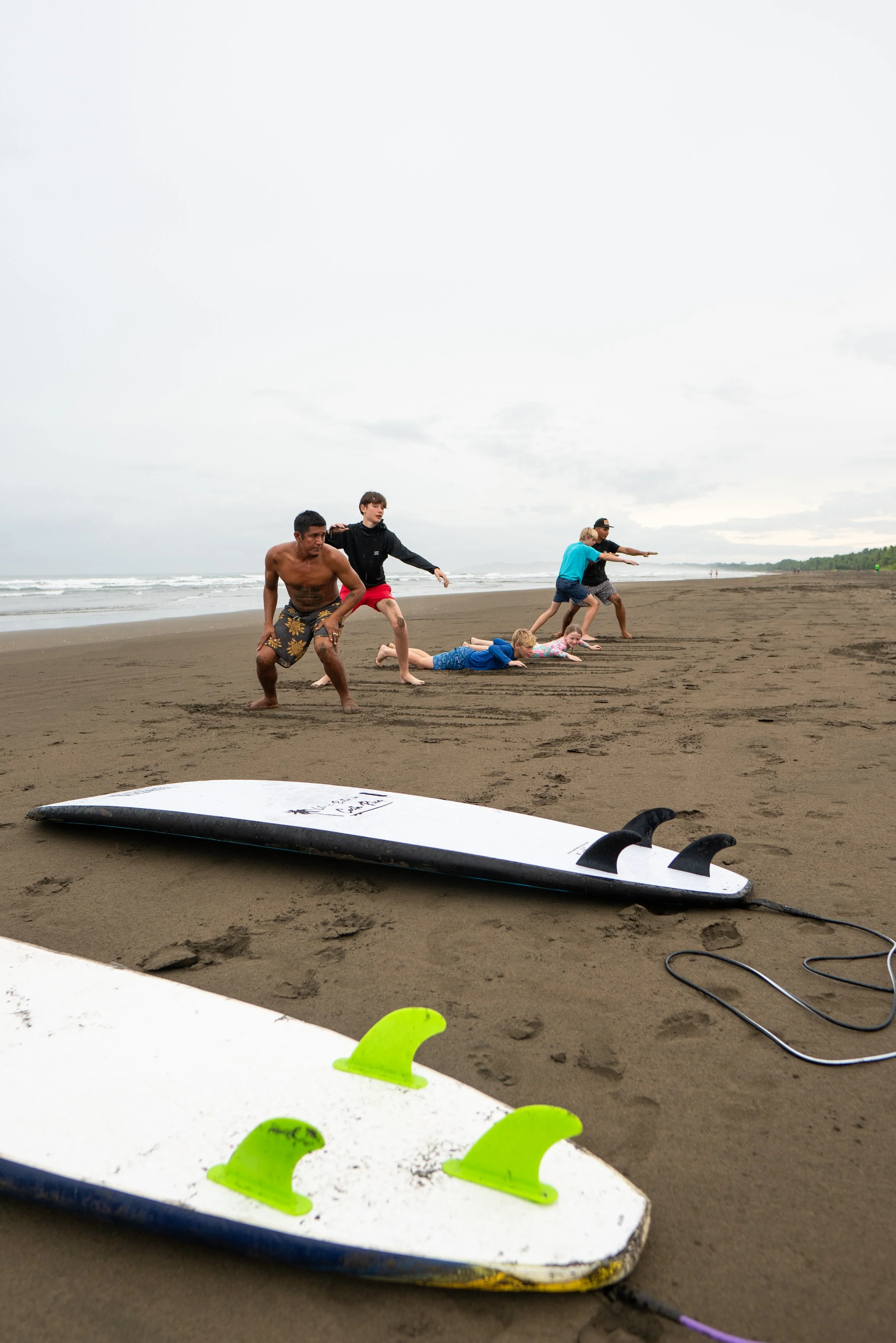 People on a beach preparing to surf, with a surfboard and fins in the foreground, and a group of children and adults in the background, some lying on the sand and others standing or crouching.