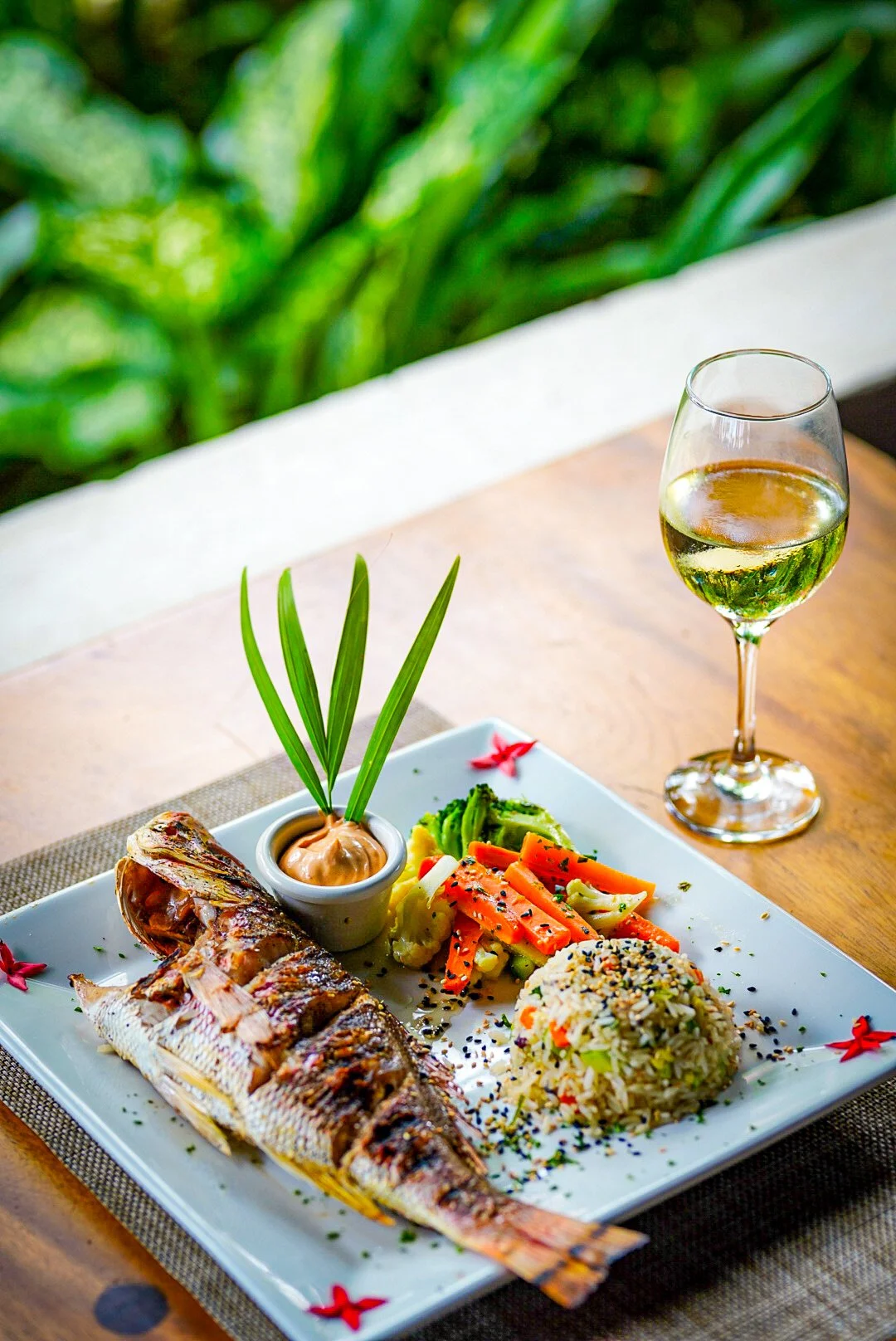 A white square plate with a whole grilled fish, vegetables, rice, and a dipping sauce, garnished with green onions. There is a glass of white wine and a small decorative flower on a wooden table with a blurred green background.