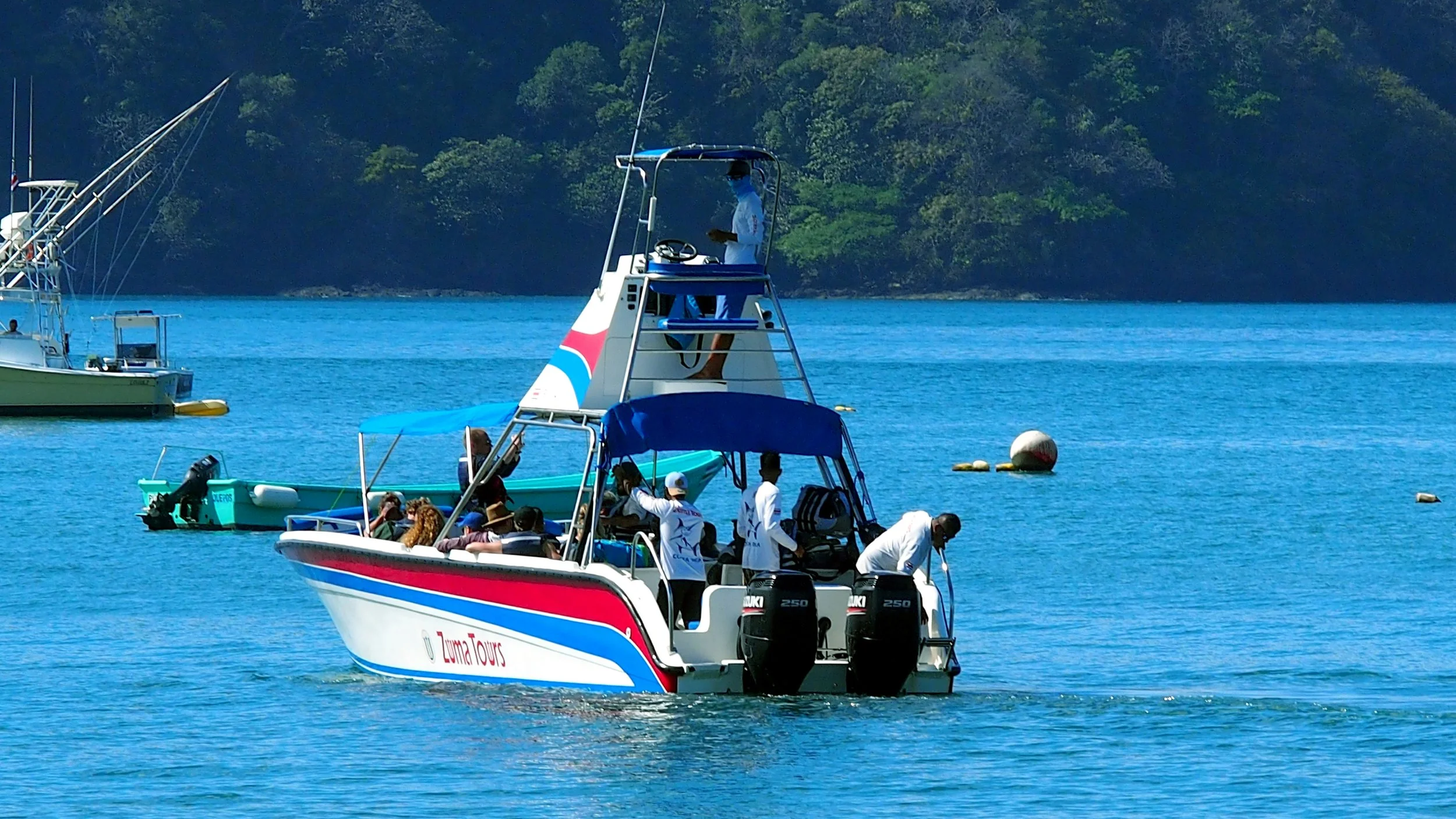 Tour boat named 'Zuma Tours' with passengers on board, two outboard motors at the back, on a calm blue body of water with boats and a green forested shoreline in the background.