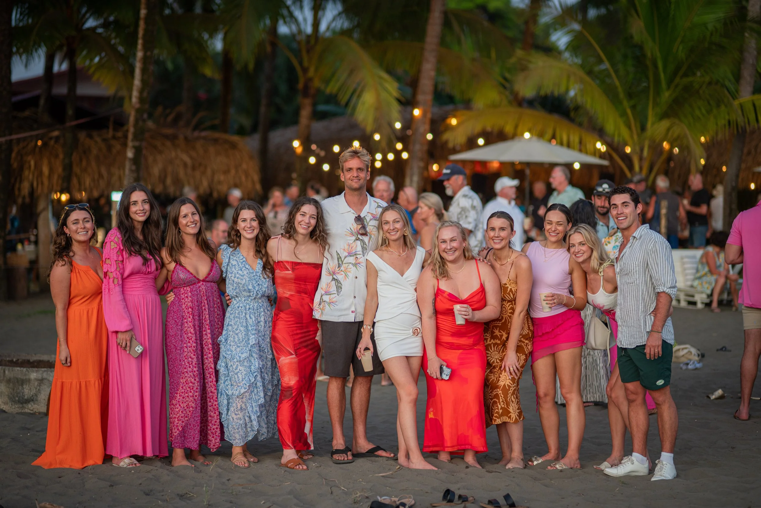 Group of people on a beach at sunset, smiling and posing for a photo, with palm trees, string lights, and beach huts in the background.