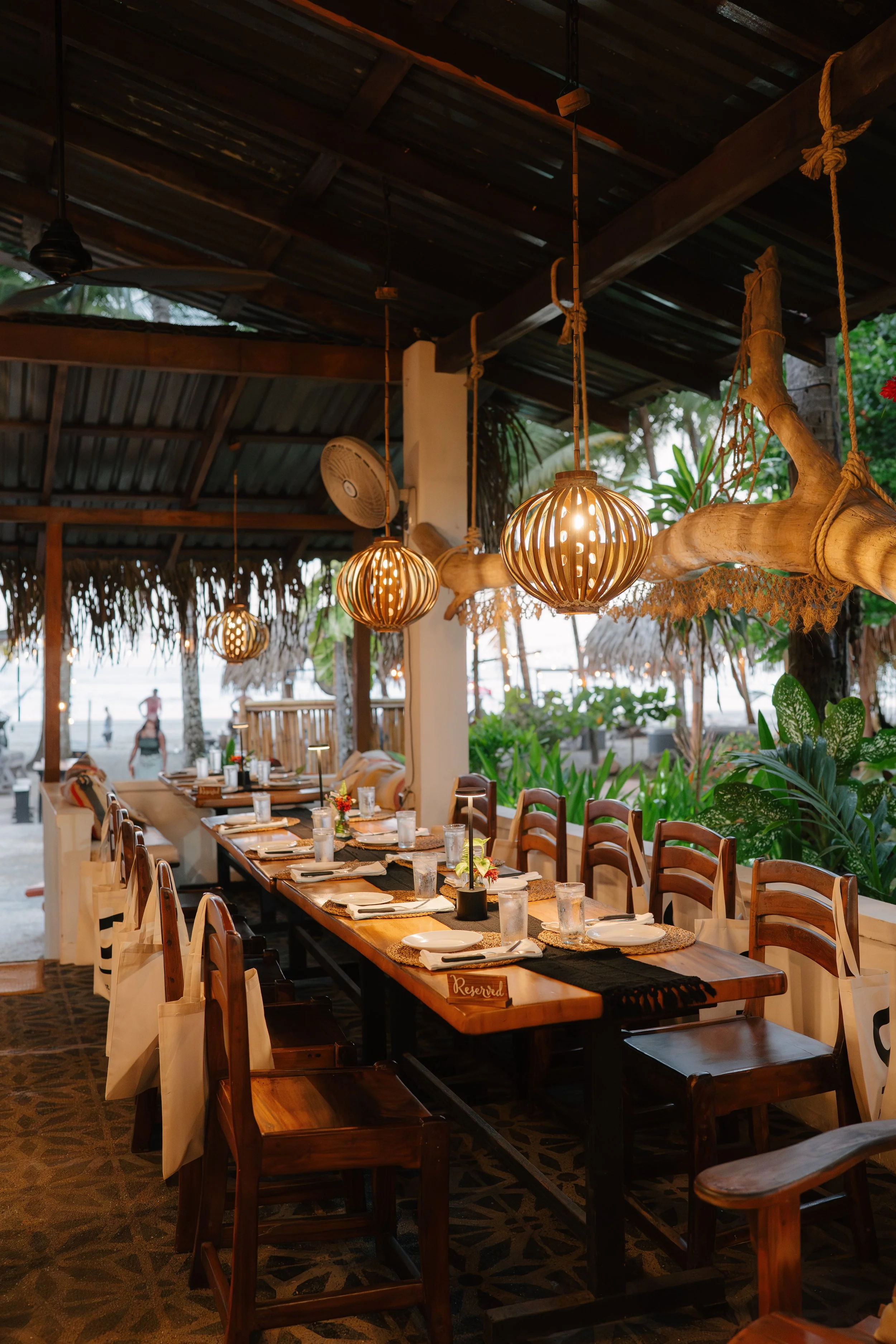A long outdoor dining table set for a meal at a seaside restaurant, with white plates, glasses, napkins, and floral centerpieces, shaded by a thatched roof and decorated with wooden hanging lantern lights, surrounded by greenery and tropical foliage.