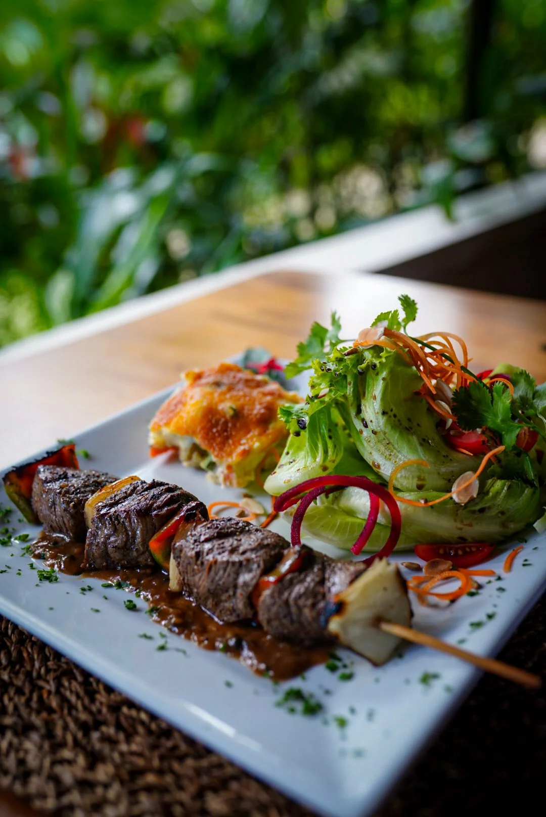Grilled beef skewers, a baked potato with cheese, and a fresh salad with lettuce, cilantro, carrots, and tomatoes on a white plate.
