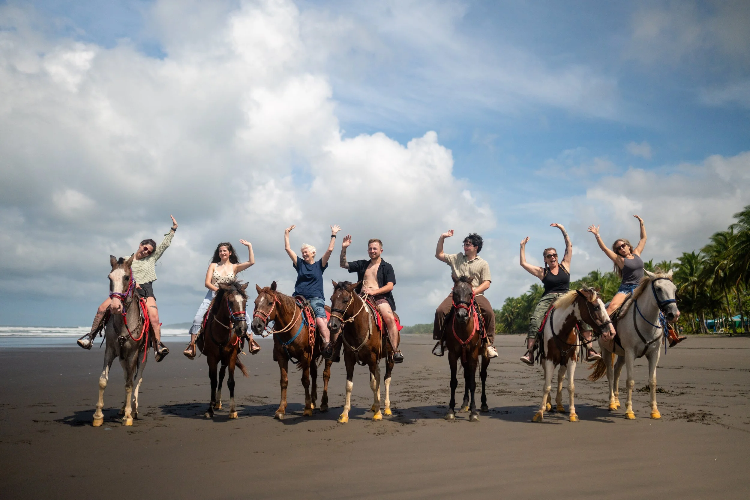 Group of seven people riding horses on a beach, smiling and waving, with cloudy sky and palm trees in the background.