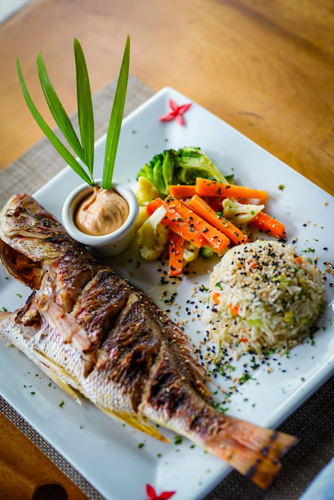 Grilled fish served with rice, mixed vegetables, and a dipping sauce on a white plate.