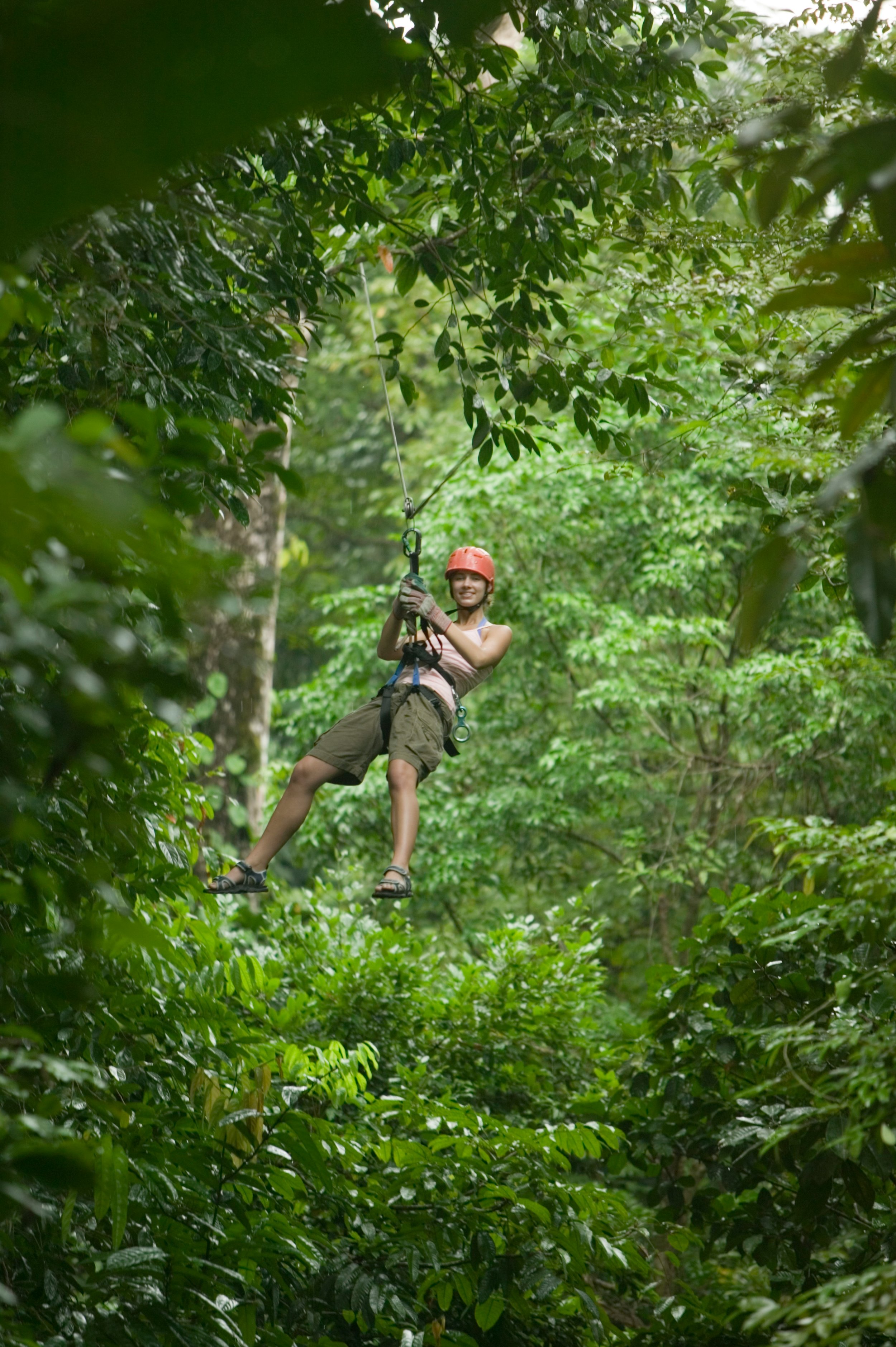 Person wearing safety helmet and harness ziplining through a dense green forest.